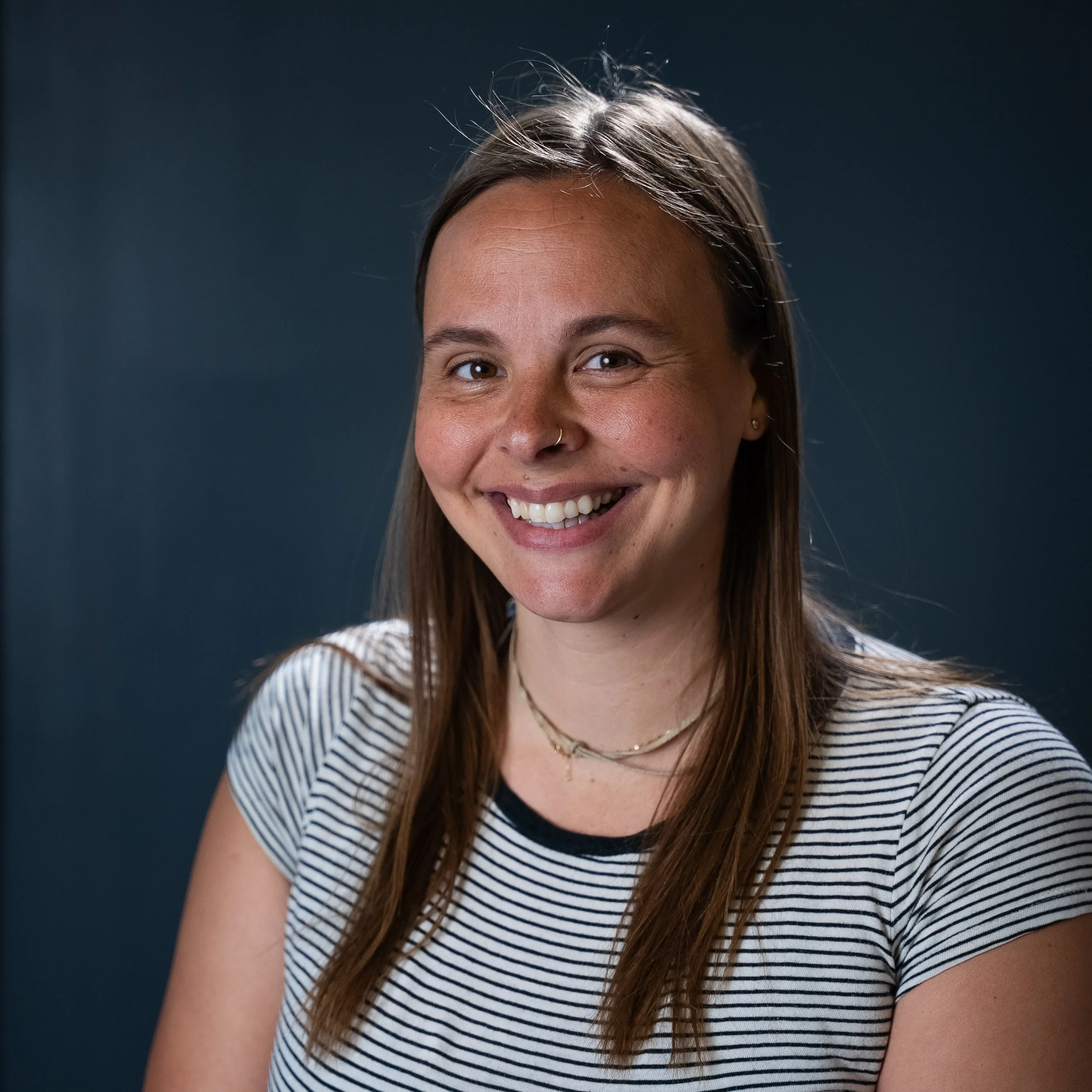 Close-up of a smiling woman with long brown hair, wearing a black and white striped shirt, standing against a dark background.