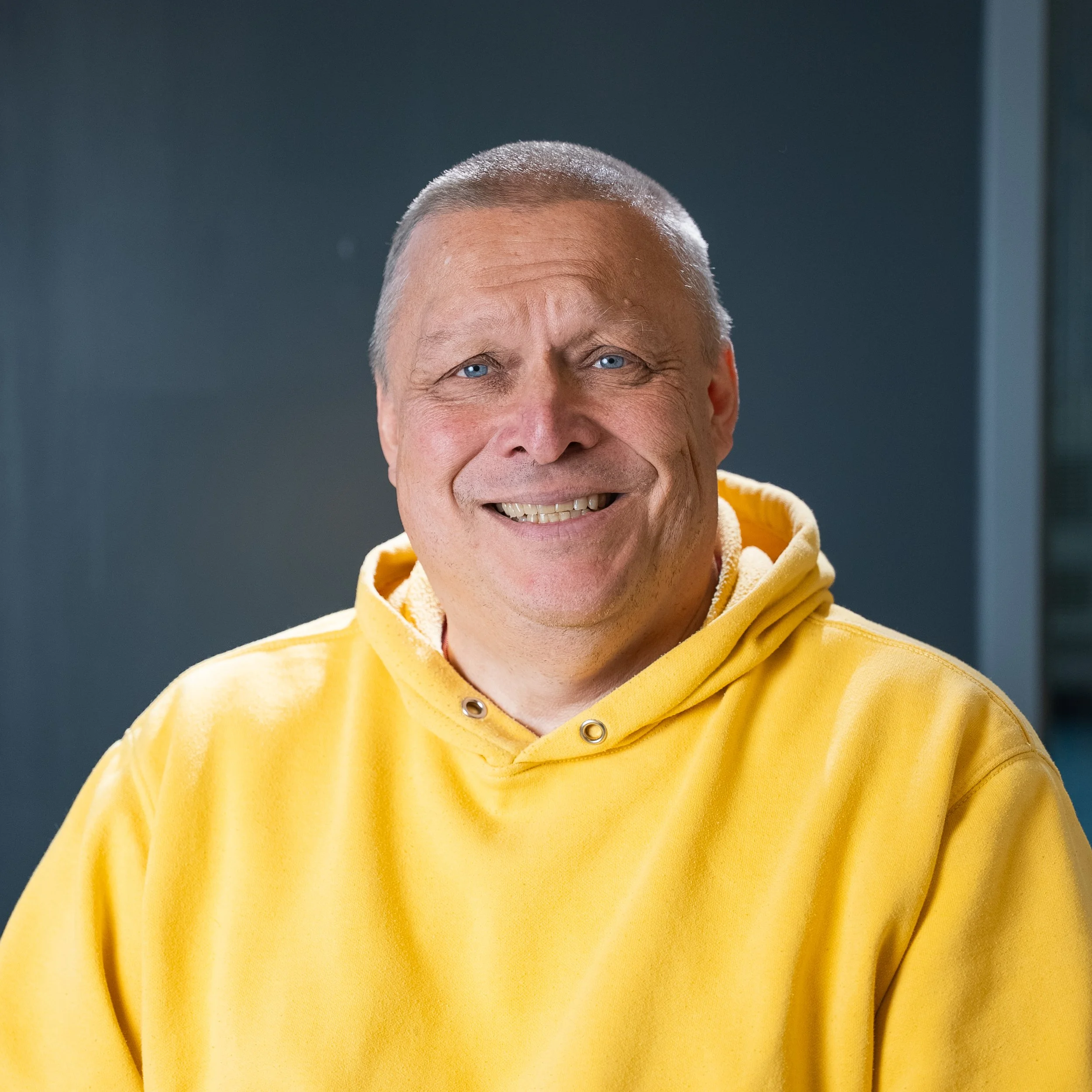 Close-up of an older man with blue eyes, short gray hair, smiling, wearing a yellow hoodie.