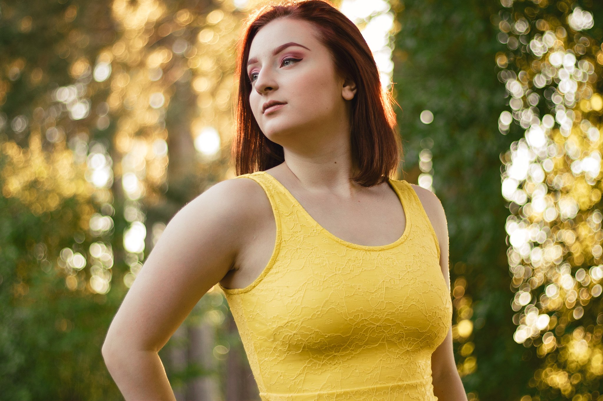 A woman with shoulder-length auburn hair and fair skin wearing a yellow sleeveless lace dress, standing outdoors in front of trees with sunlight creating a bokeh background.
