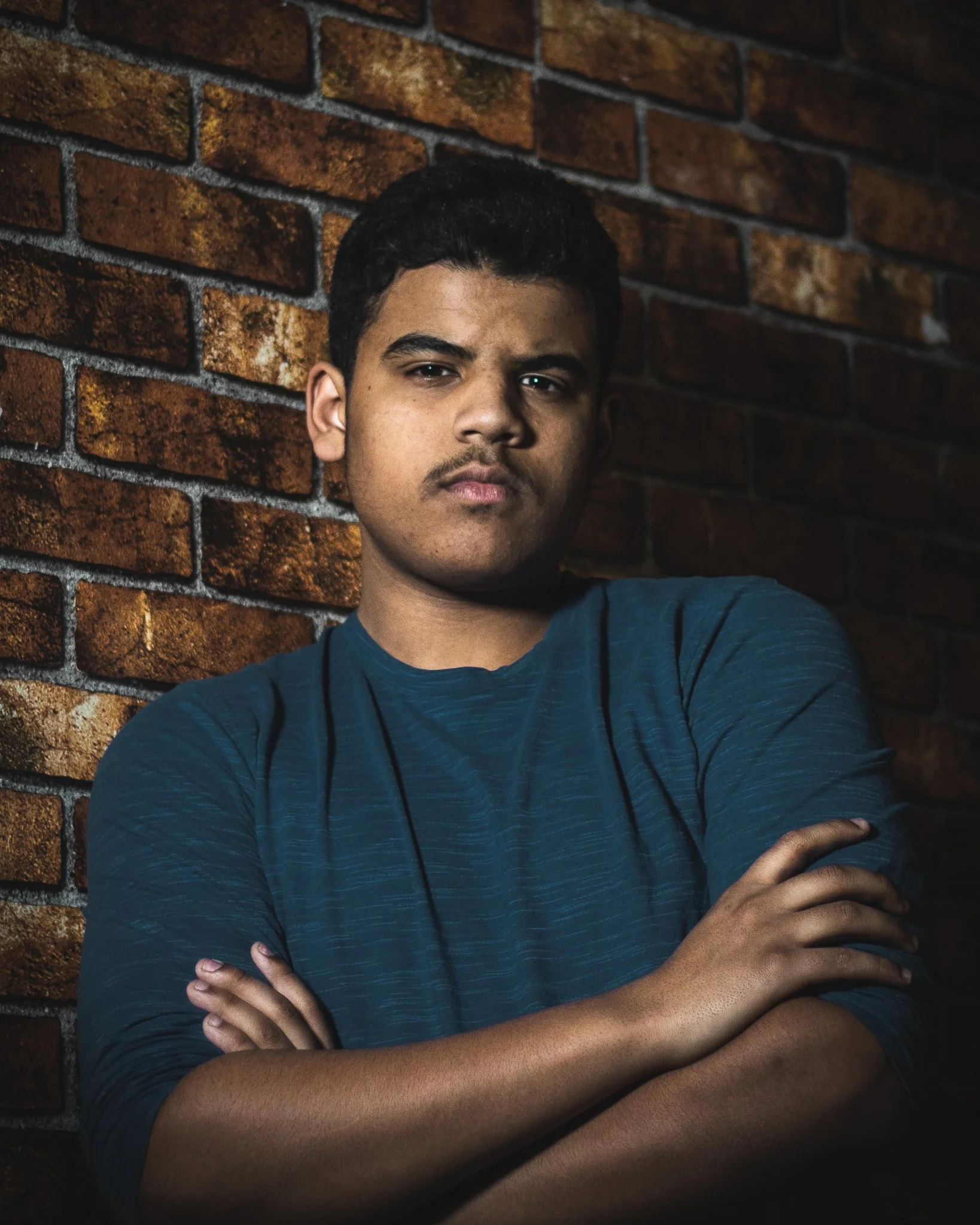 A young man with short black hair and brown skin, wearing a dark blue shirt, standing against a brick wall. He has a serious expression and crossed arms.