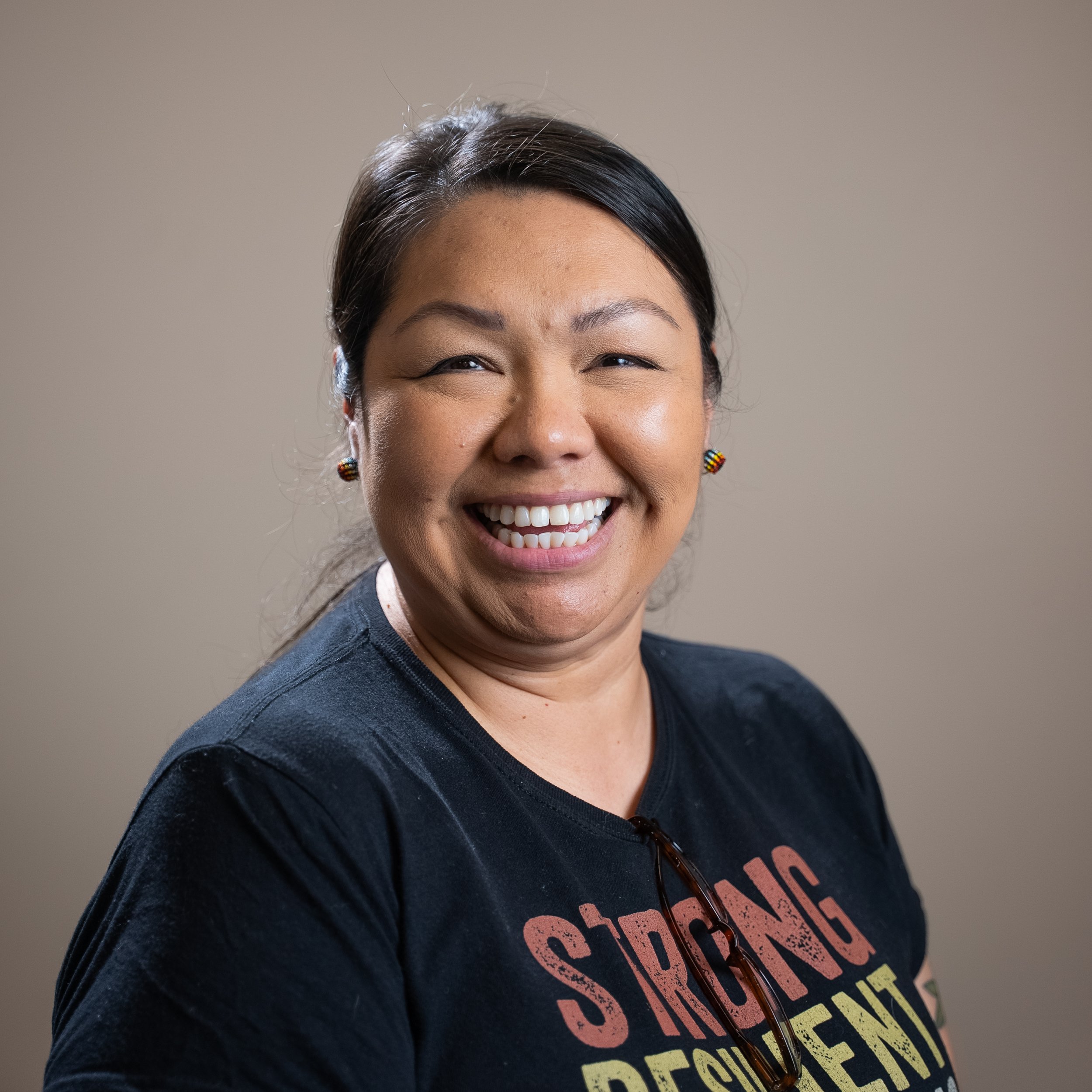 Close-up of a smiling woman with dark hair, earrings, and wearing a dark shirt with colorful text, against a plain background.