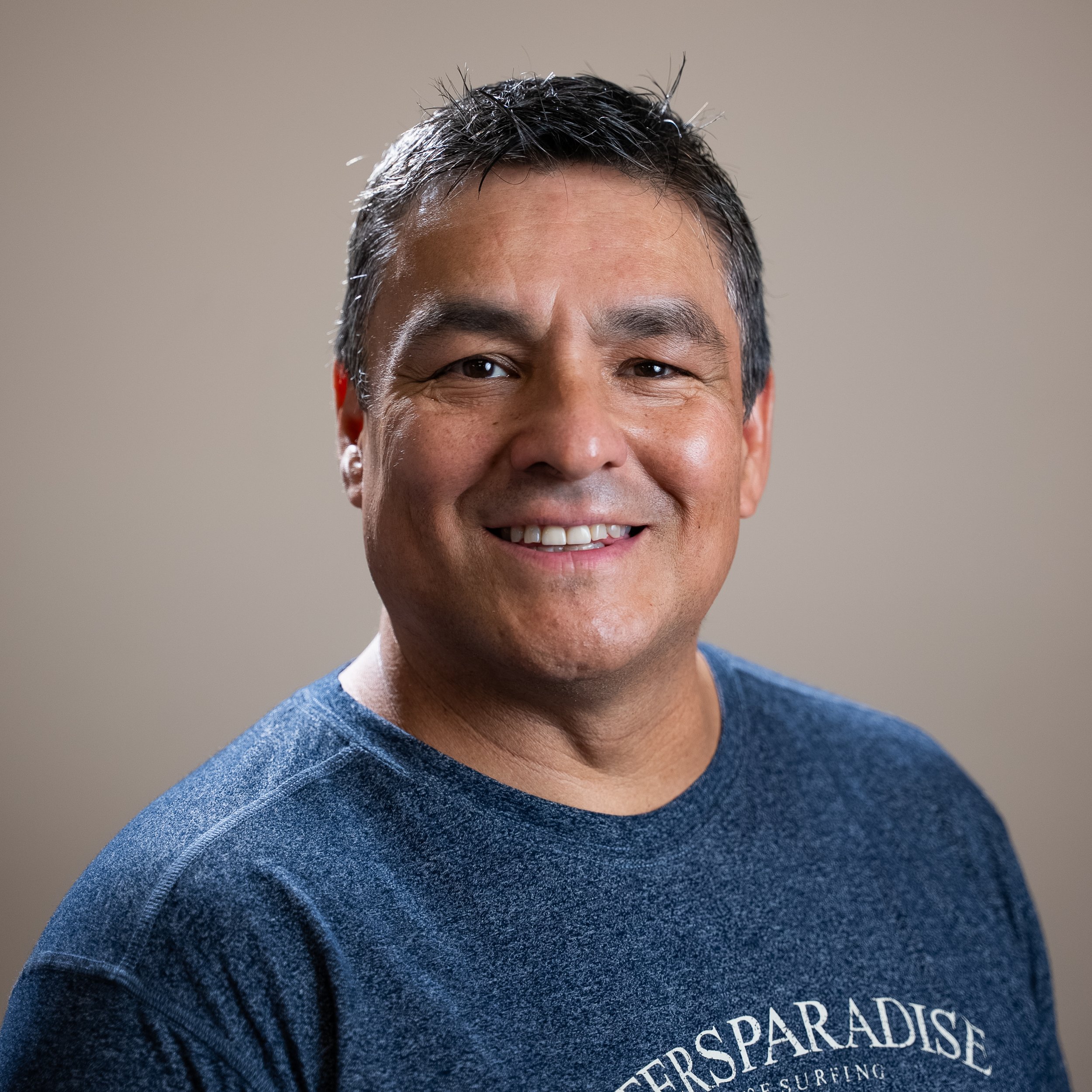 Close-up of a smiling man with short dark hair, wearing a blue sweatshirt with white text that reads 'PARADISE' and other partially visible words, against a neutral background.