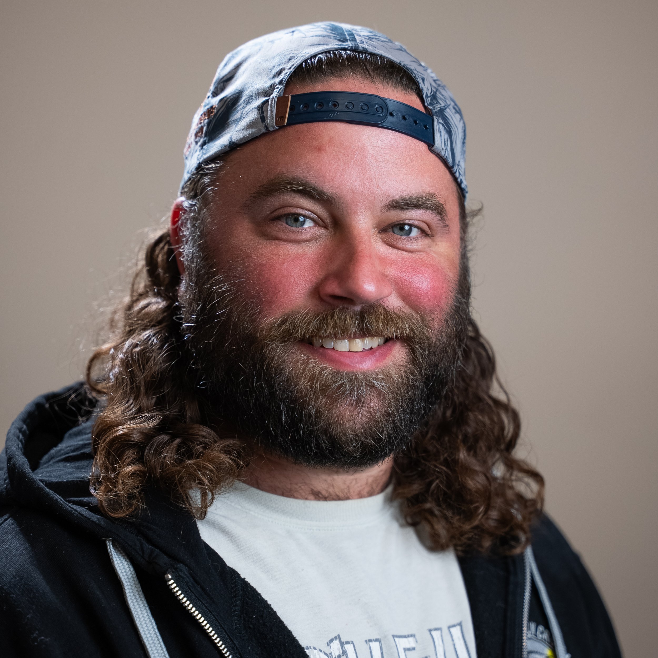 Close-up of a man with long, curly hair, a beard, wearing a backwards baseball cap, a white T-shirt, and a black zip-up hoodie, smiling.