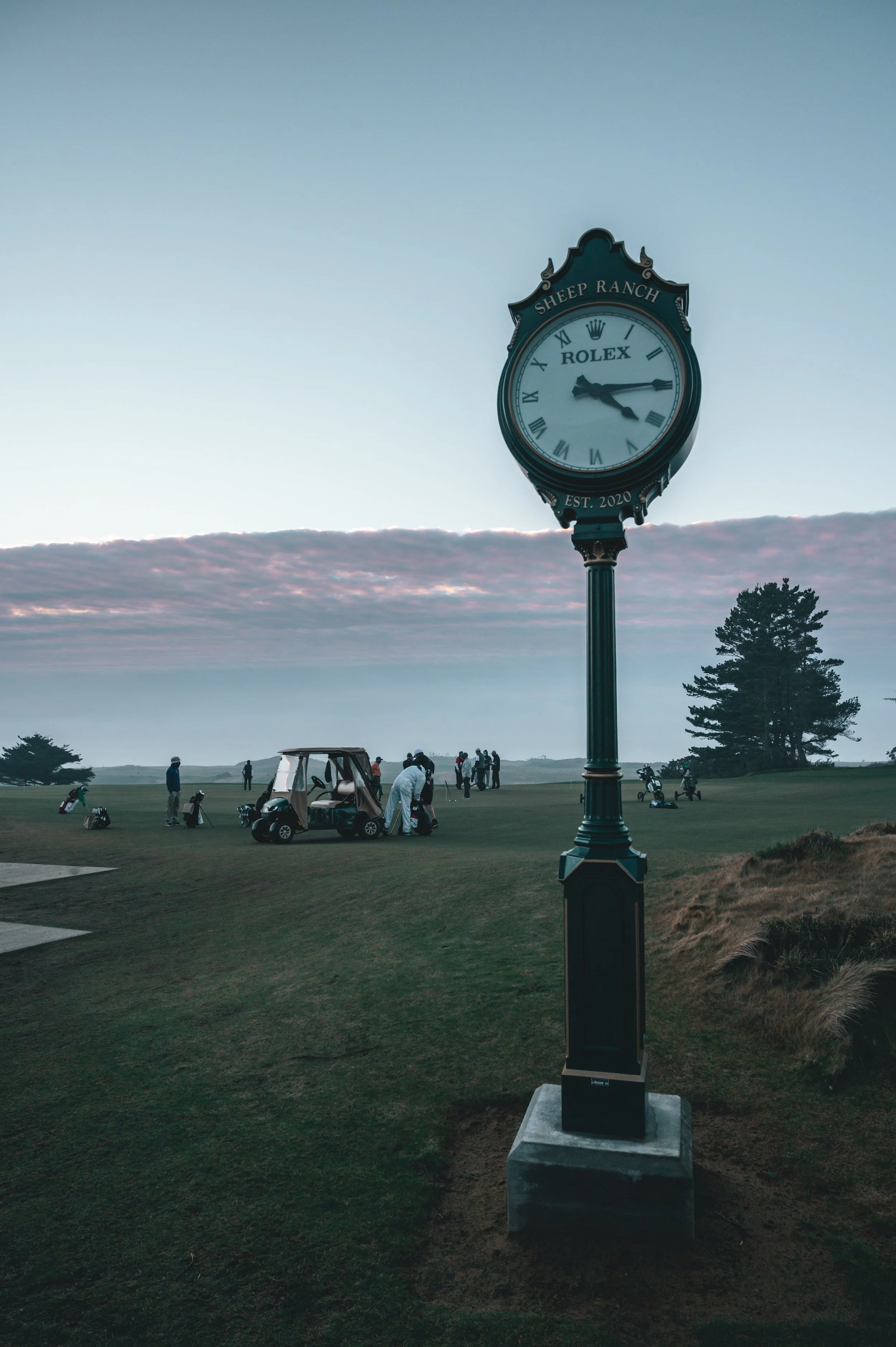 Golf course with people playing, a golf cart, and a tall decorative clock with the inscription "Sheep Ranch" and "Rolex" in the foreground, during dusk or dawn.
