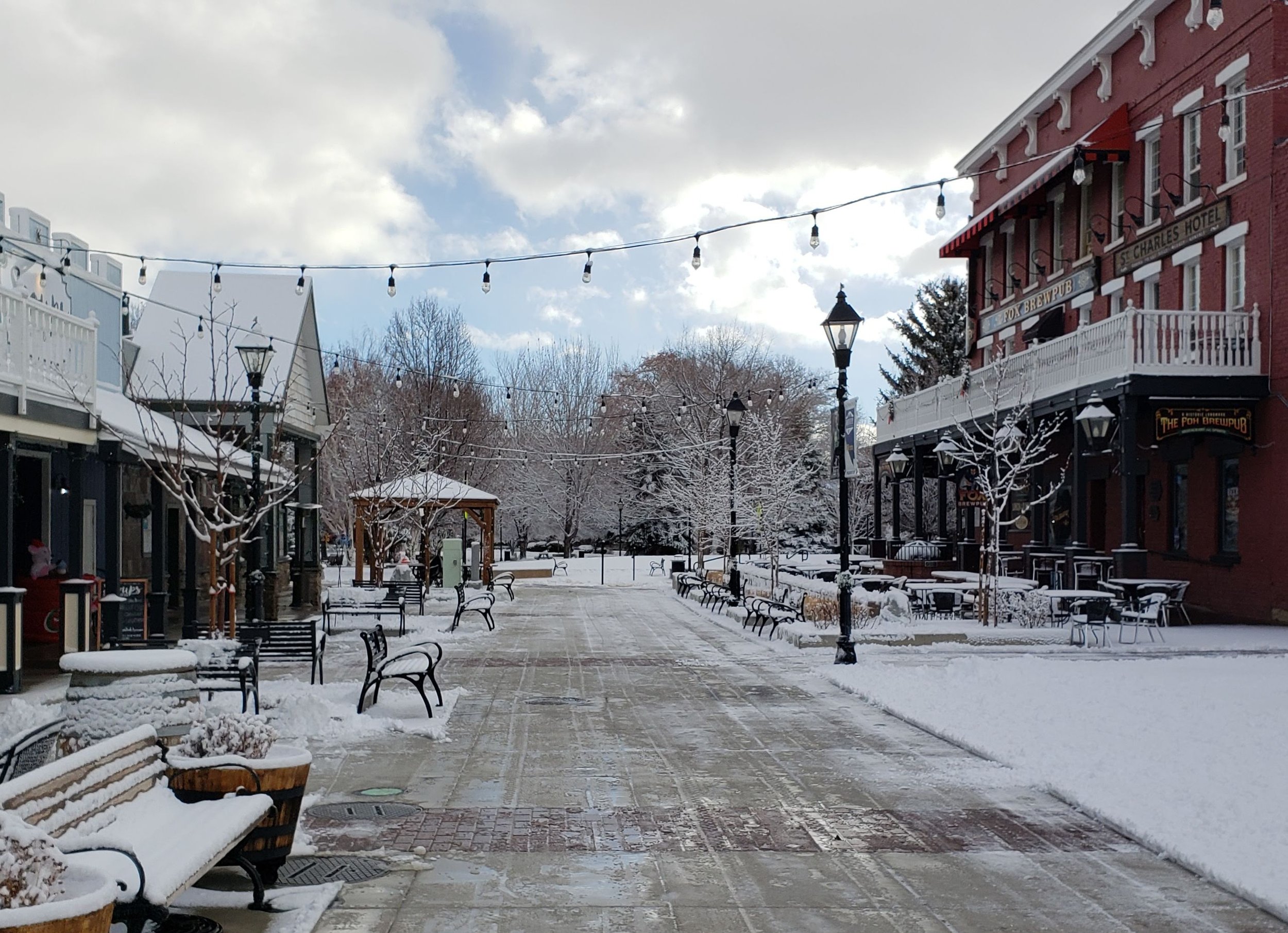 A snow-covered outdoor plaza with benches, trees, and lampposts, surrounded by buildings including a red brick hotel with a balcony and string lights overhead.