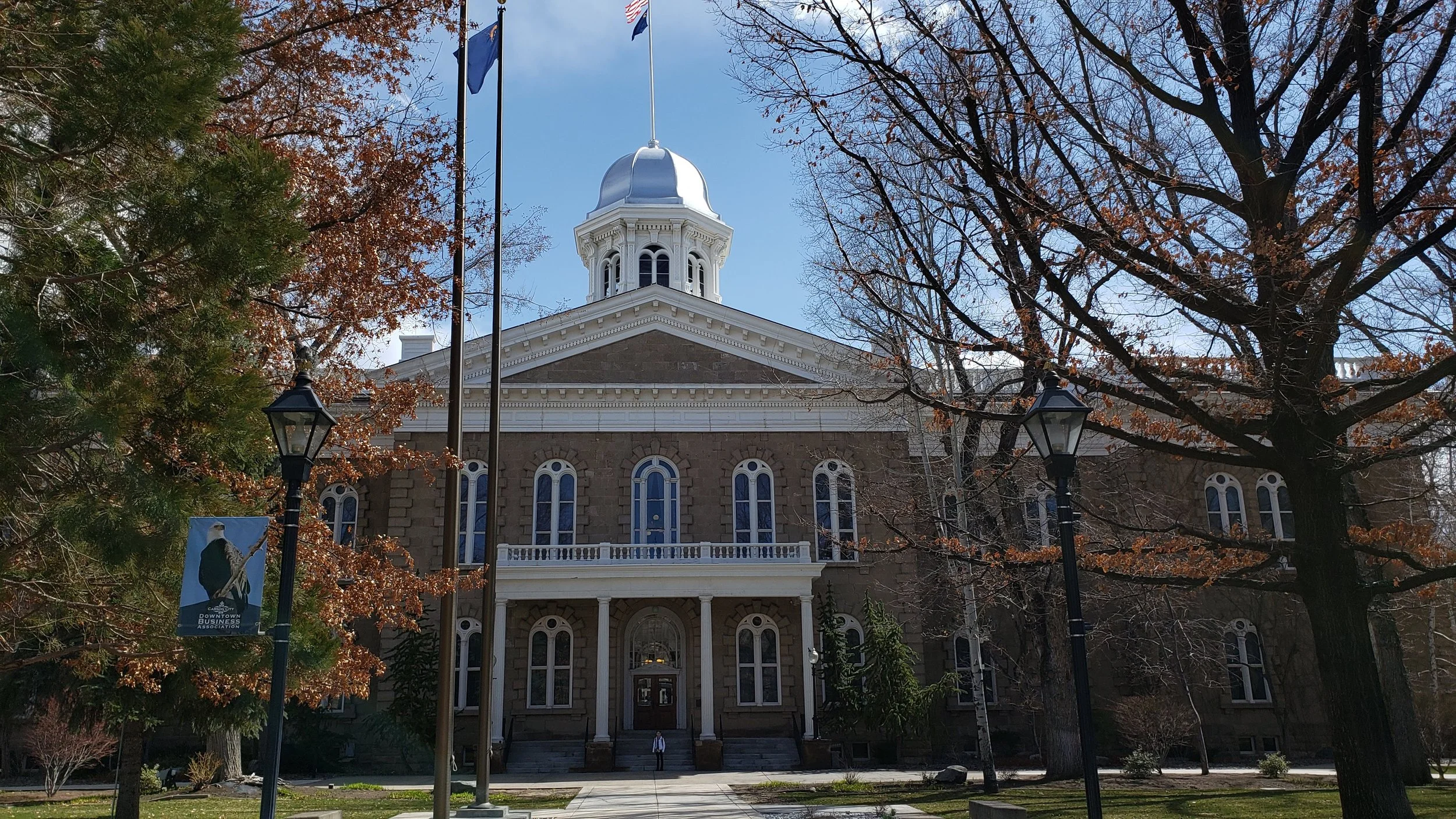 A historic stone building with a prominent clock tower and a dome, framed by trees with autumn leaves and two street lamps, under a blue sky.