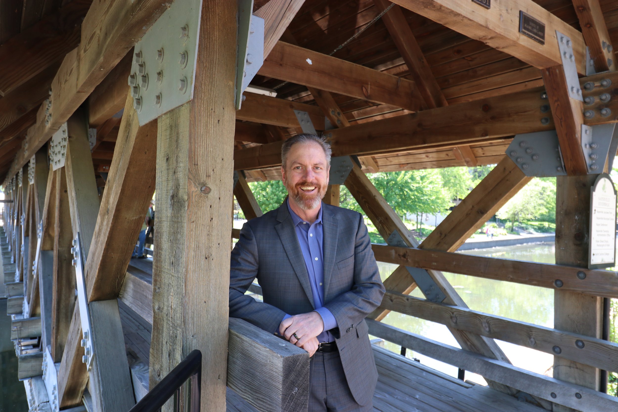 A man in a gray suit with a blue shirt is standing on a wooden bridge structure with a smile, surrounded by trees and greenery.