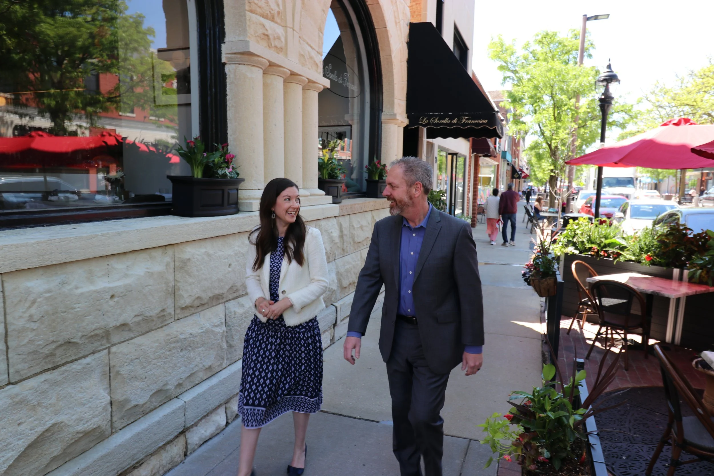 A woman and a man walking together on a city sidewalk, smiling and looking at each other. The woman is wearing a white cardigan and a blue printed dress, and the man is dressed in a dark suit with a blue shirt. The background shows a stone building with large windows, potted plants, and pedestrians walking by, with trees, outdoor tables, and umbrellas.