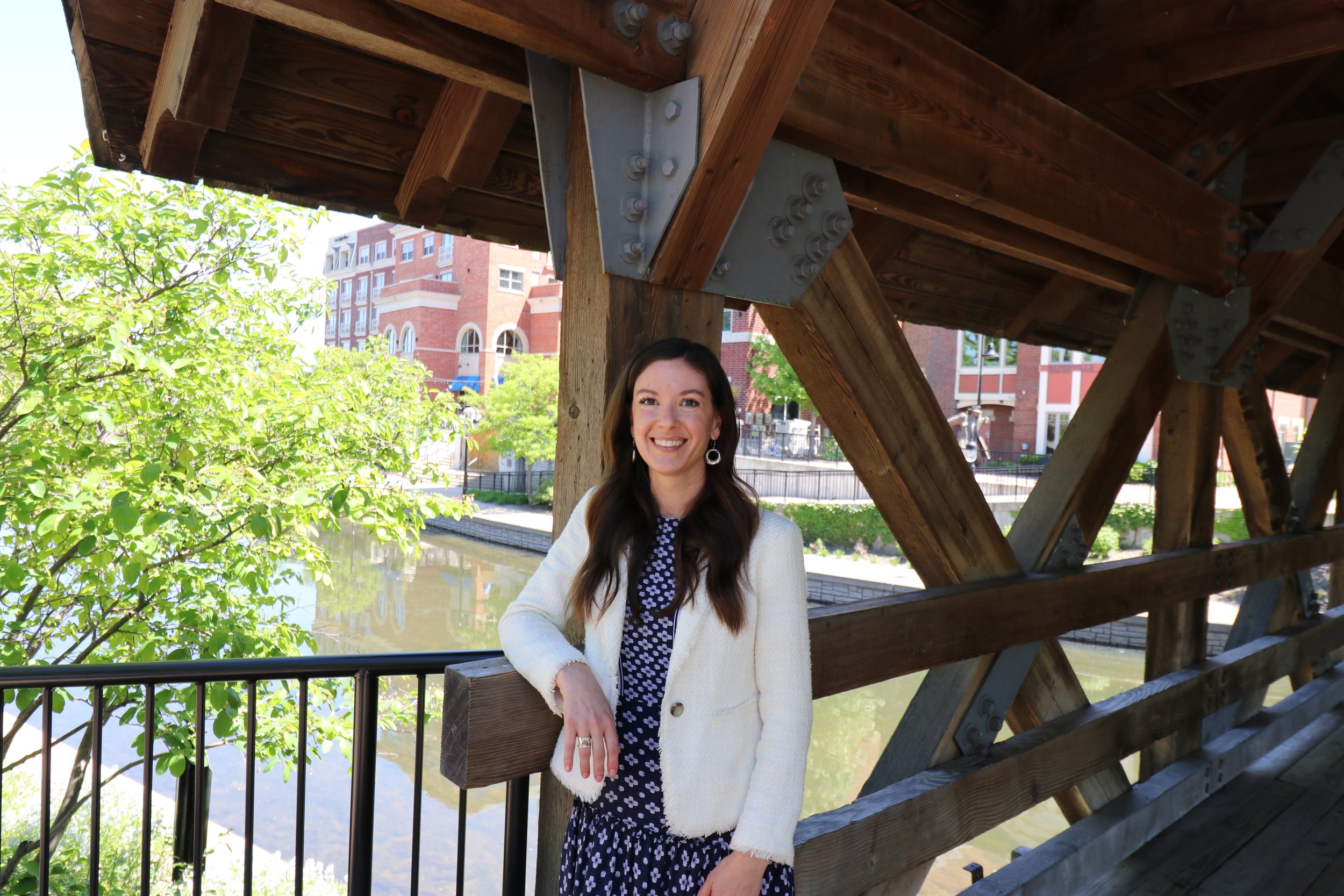 A woman with long dark hair, wearing a white cardigan and a black dress with white polka dots, smiling and leaning on a railing under a wooden bridge near a pond, with trees and red-brick buildings in the background.
