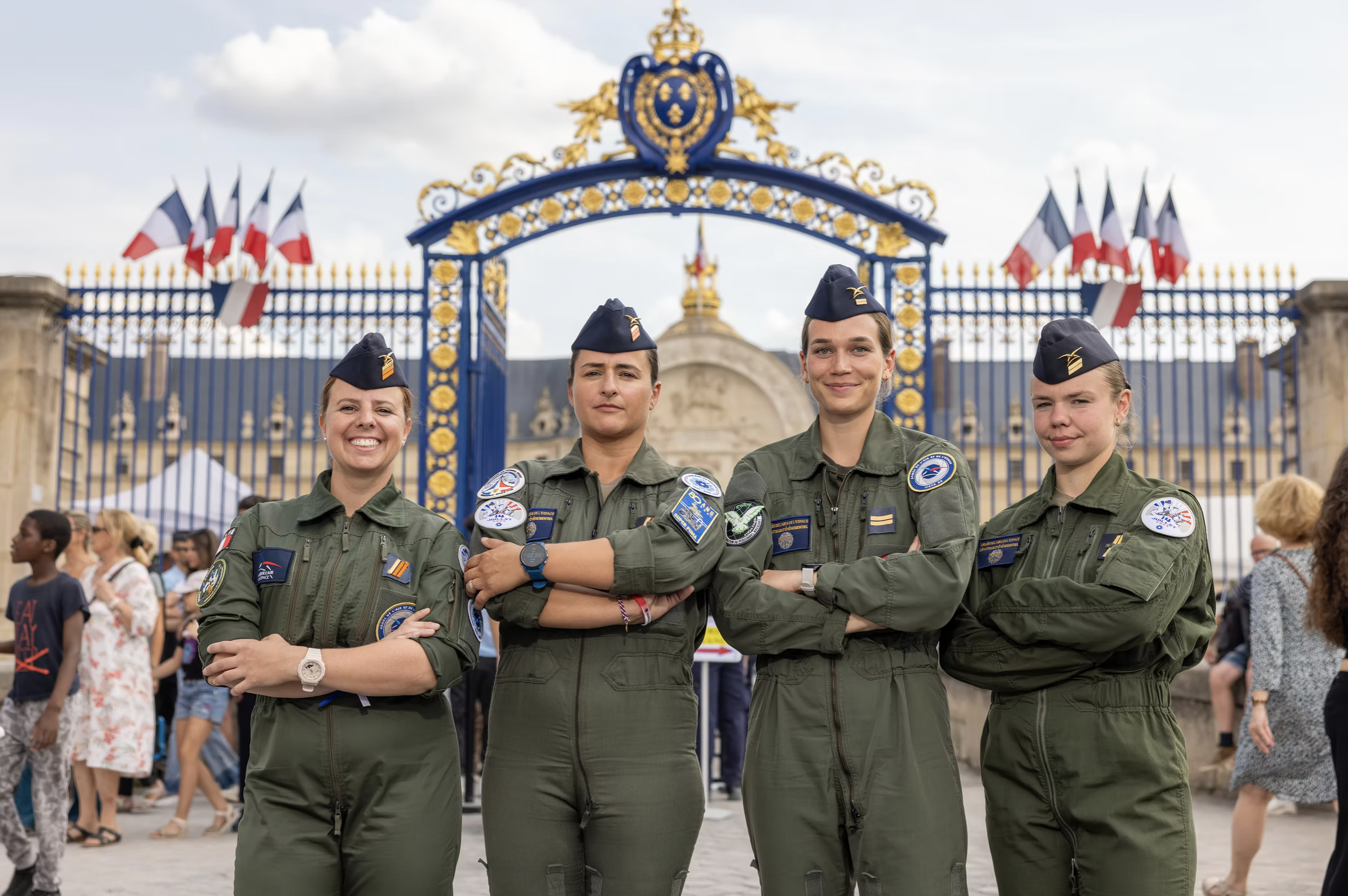 Quatre femmes en uniformes militaires se tiennent côte à côte avec leurs bras croisés devant une entrée ornée de drapeaux français, souriant vers l'appareil photo.