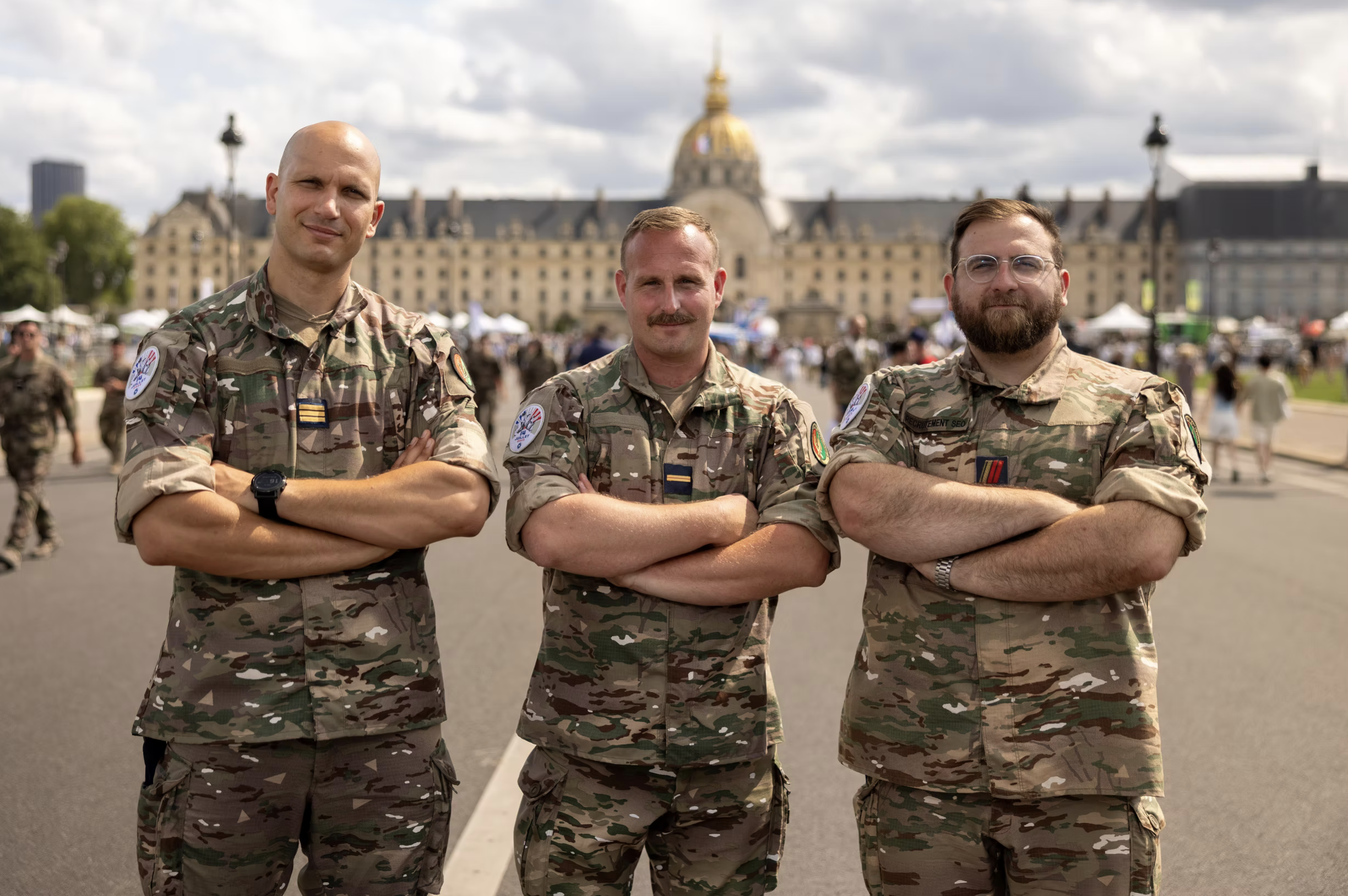 Trois soldats en uniforme camouflage posent avec leurs bras croisés devant un bâtiment célèbre dans un parc public.