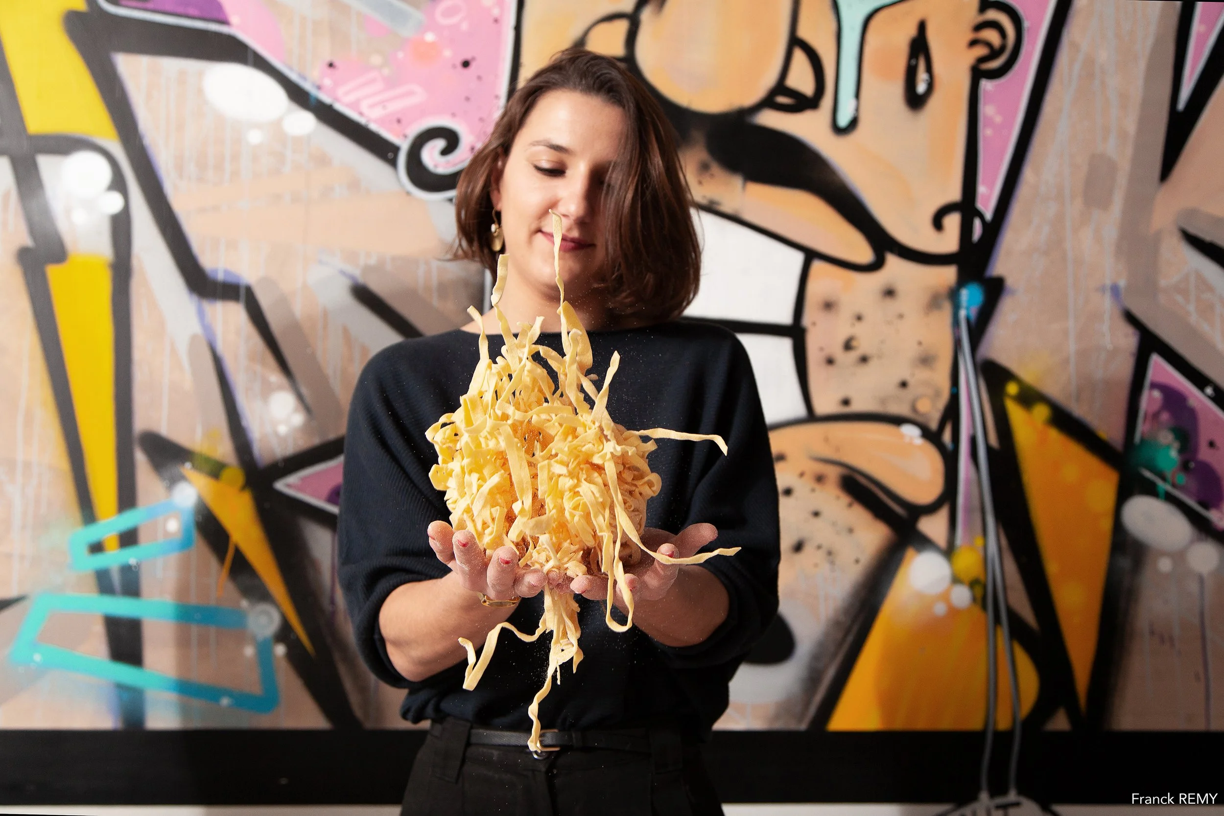 Une femme tenant une poignée de pâtes ou de fromage râpé, dans un décor de graffiti coloré en arrière-plan.