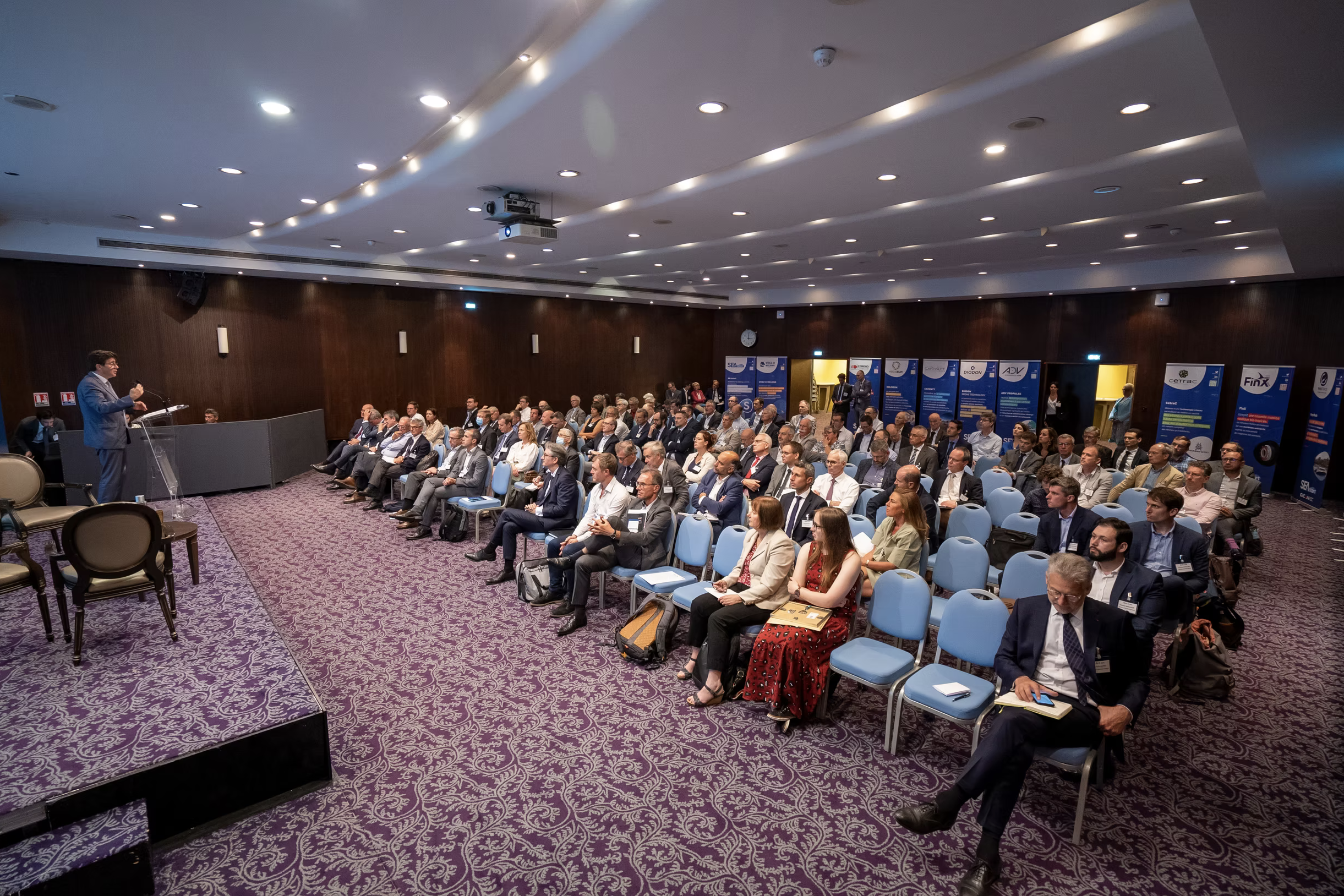 Conférence en salle avec un orateur debout à une rampe, face à un public nombreux assis dans des sièges bleus, dans une salle de conférence avec un plafond lumineux et un tapis violet à motif.