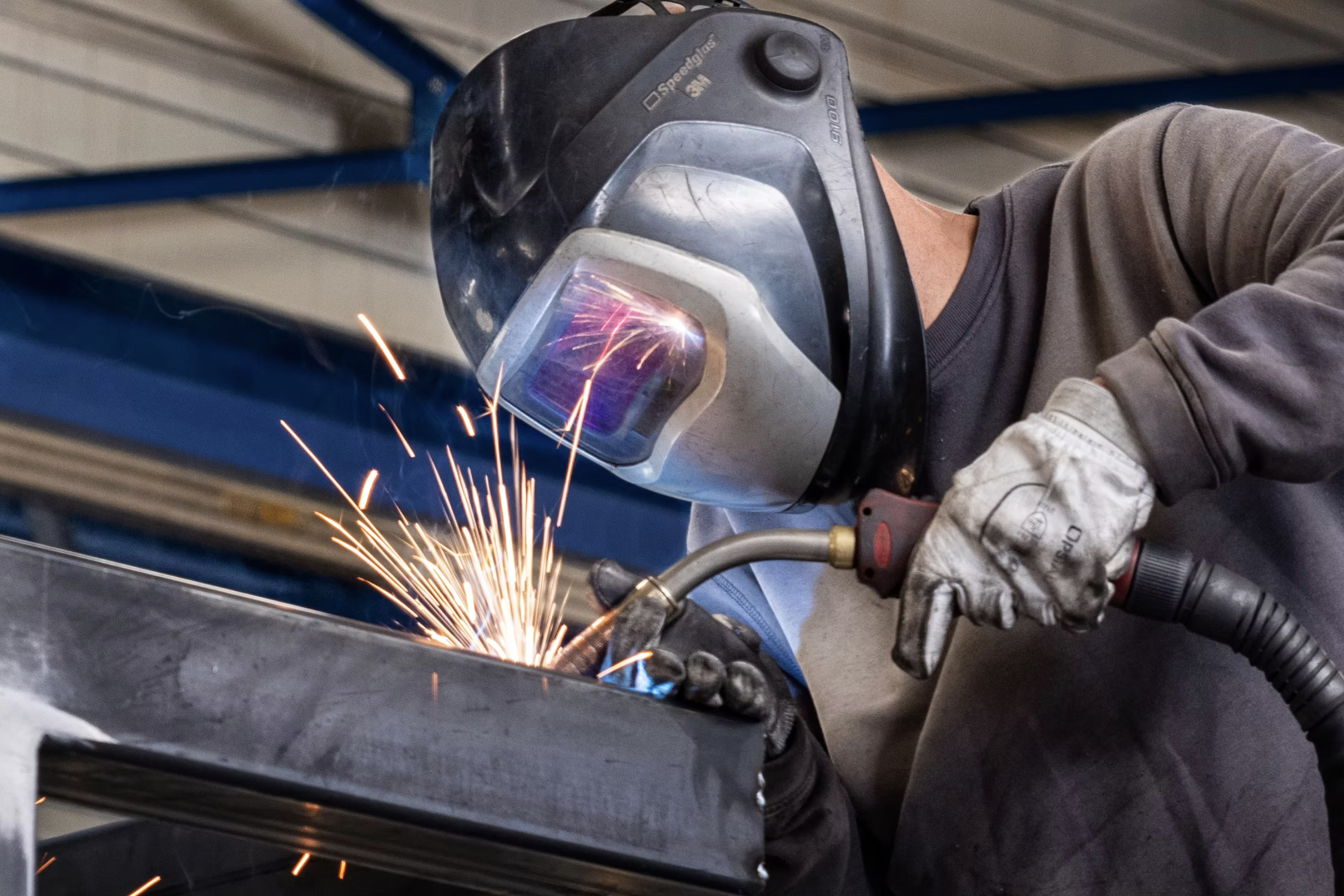 Un ouvrier portant un casque de soudage et des gants utilise une machine à souder pour réaliser une soudure sur une pièce métallique, avec des étincelles visibles.