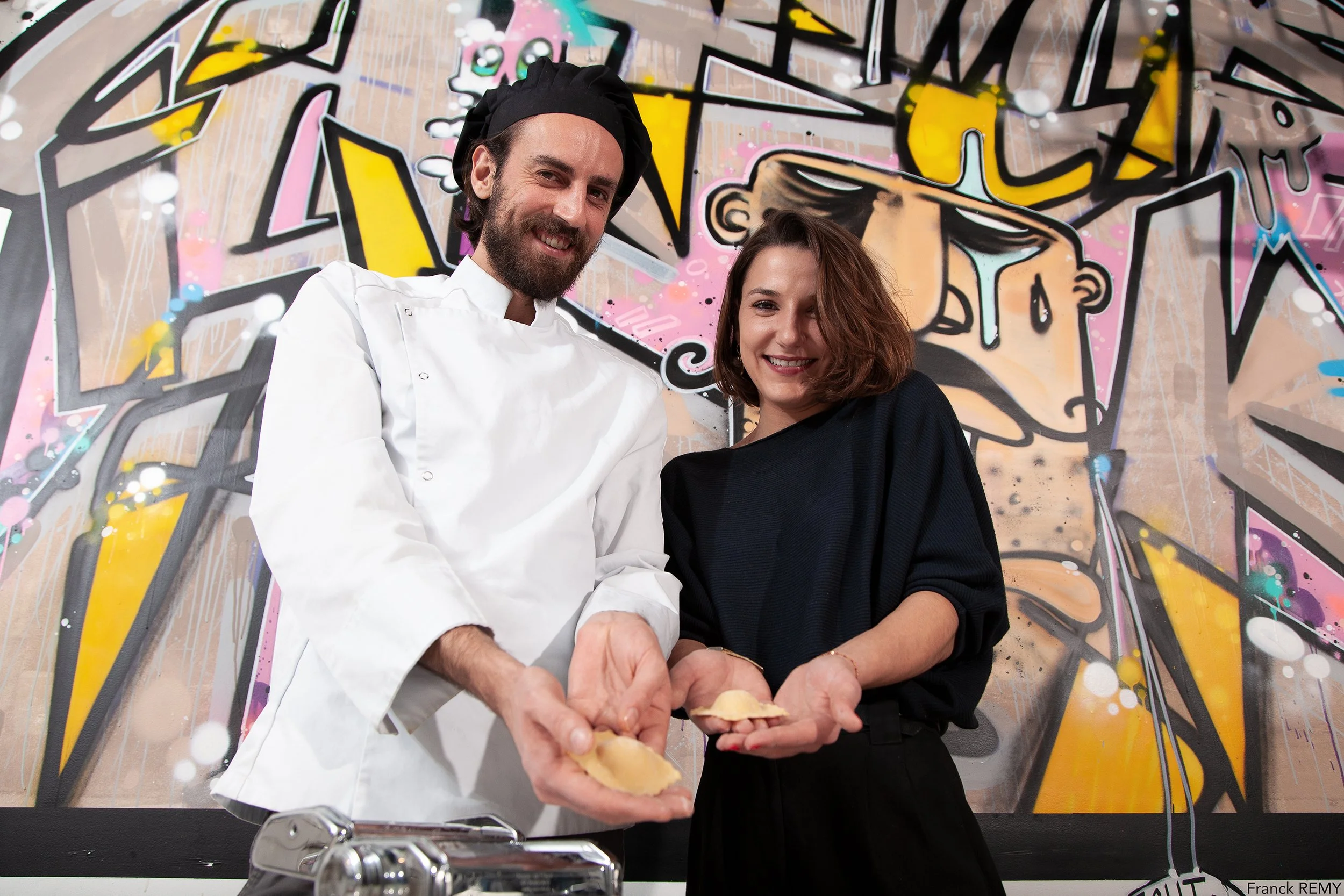 Un chef et une femme souriants montrent des raviolis faits maison, avec un graffiti coloré en arrière-plan.