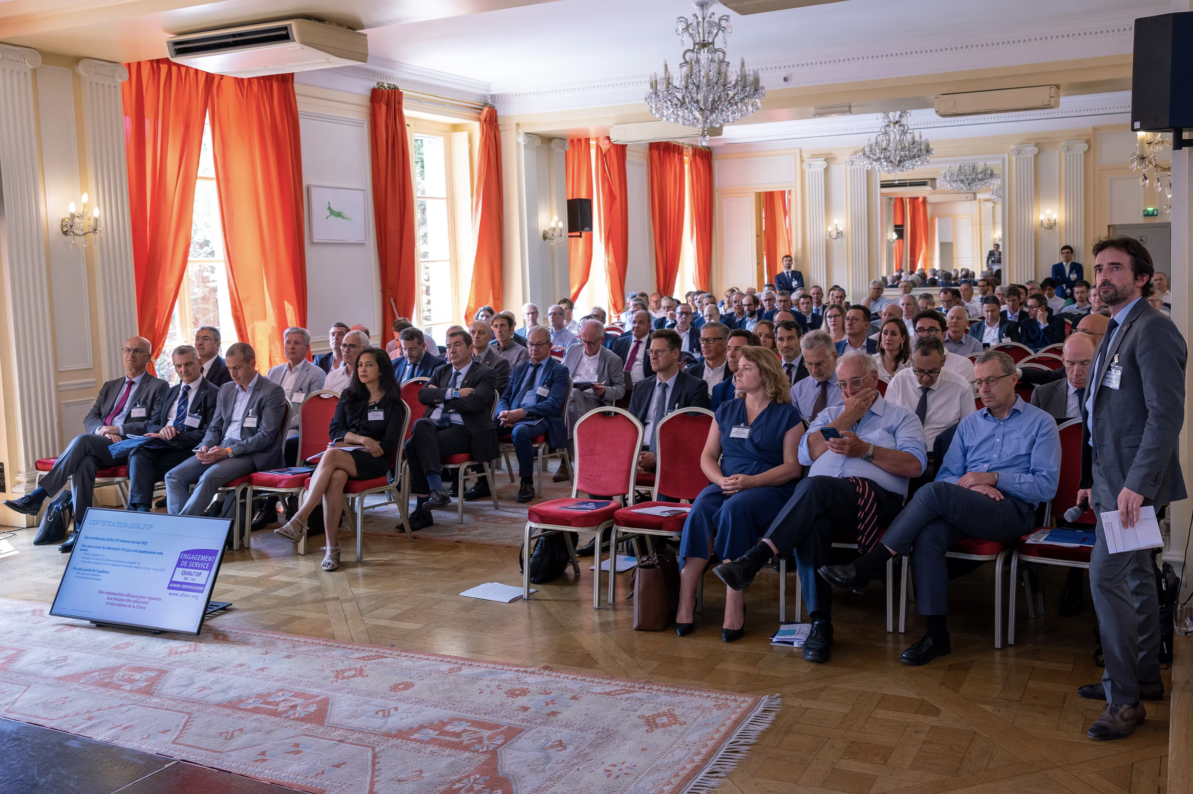 Une salle de conférence remplie de personnes assises, avec des rideaux orange, des chandeliers, et un écran à l'avant. Un homme en costume se tient à droite, tenant des papiers.