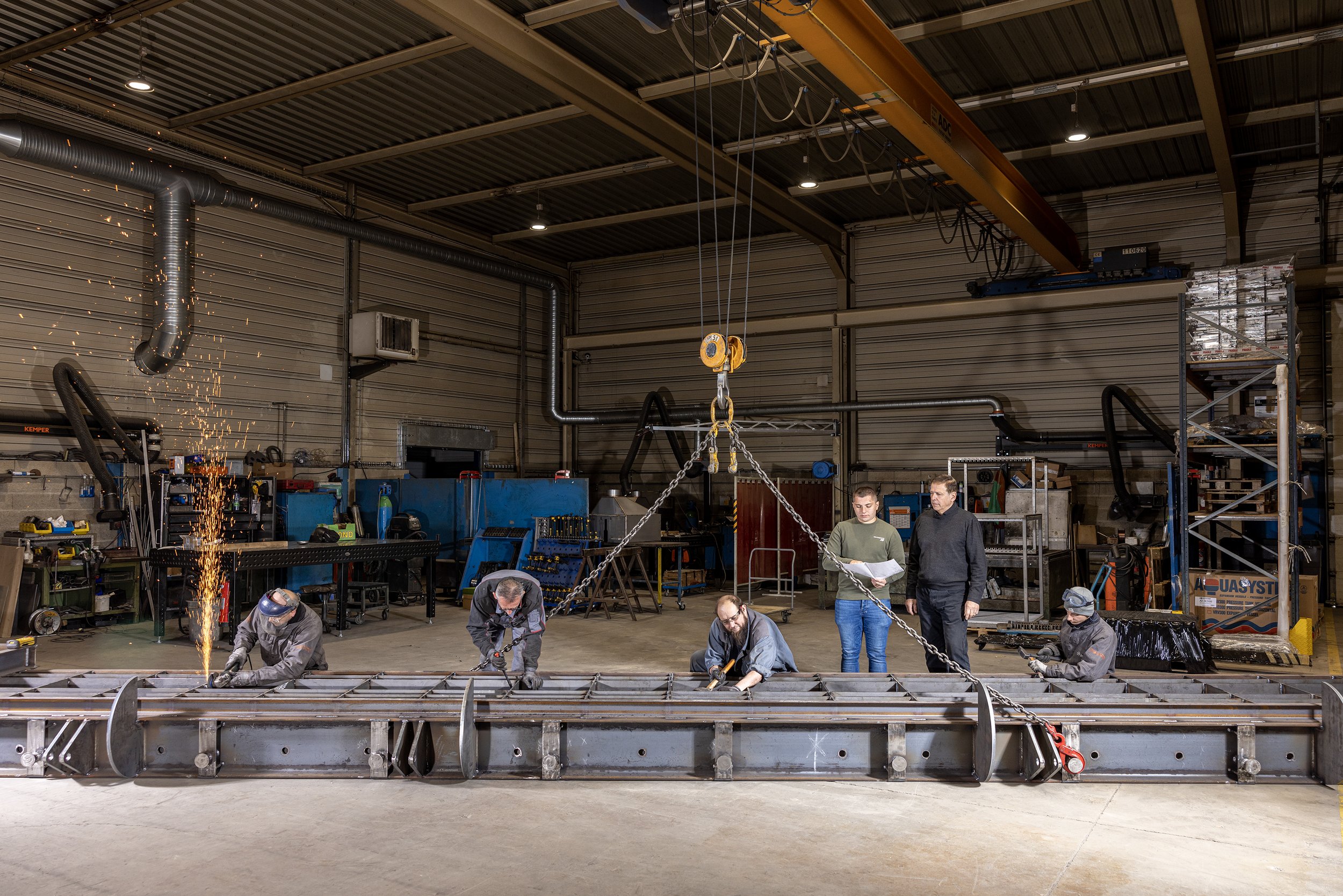 Des ouvriers travaillent sur une grande pièce métallique dans un atelier industriel, certains utilisant des outils de soudure et un cric suspendu.