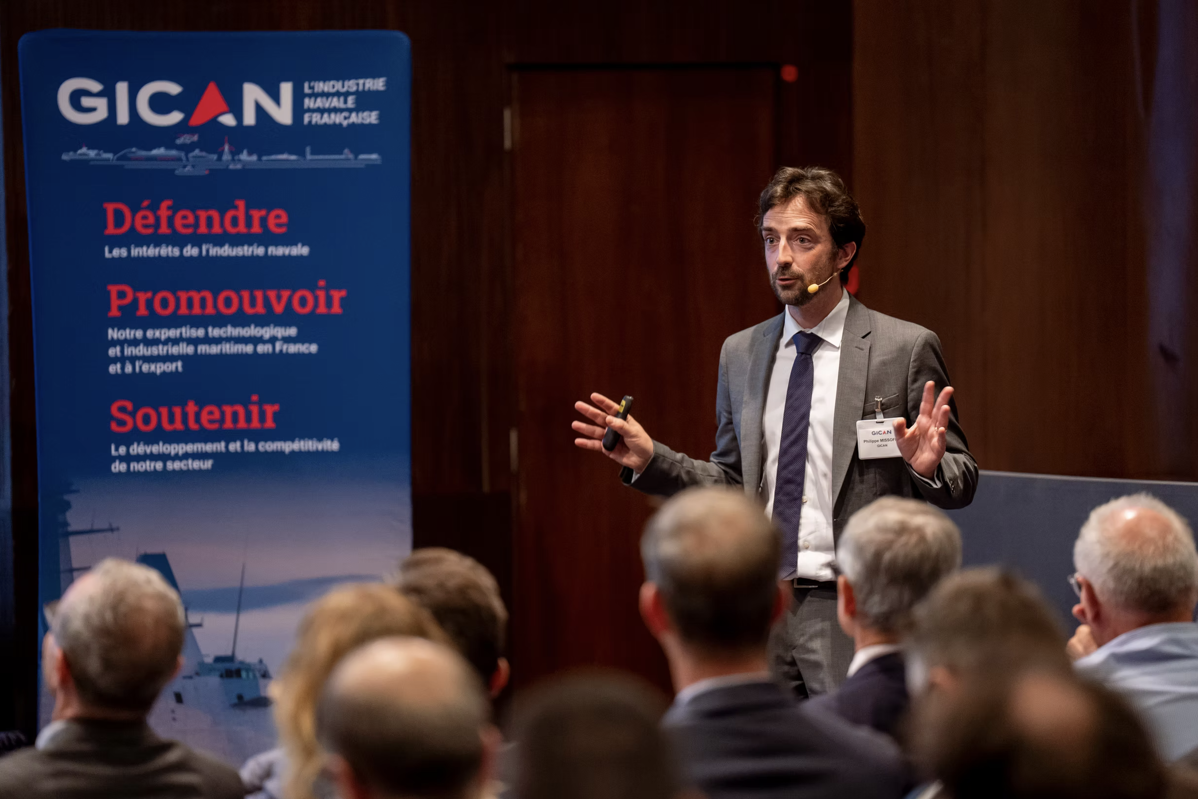 Un homme en costume gris parle lors d'une conférence, avec un public en arrière-plan, devant une bannière bleue de GICAN qui présente les objectifs de l'entreprise liés à l'industrie navale française.