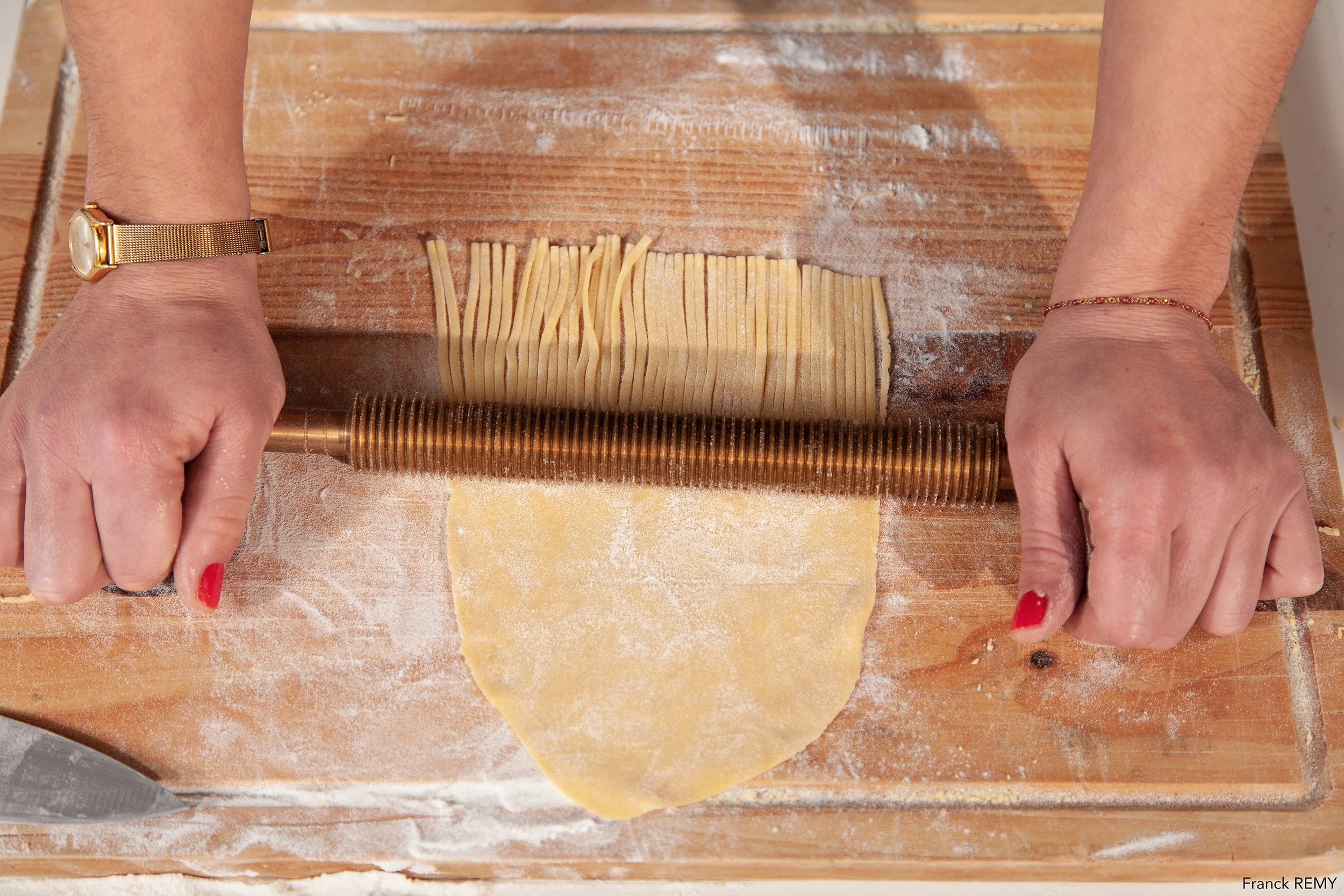Deux mains qui utilisent un rouleau à pâtisserie pour aplatir de la pâte à pâtes sur une surface en bois farinée.
