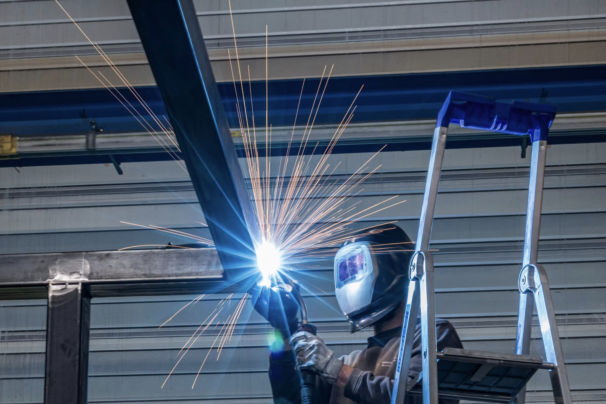Un soudeur portant un casque de protection et des gants, utilisant un poste à souder pour assembler deux pièces métalliques, avec des étincelles visibles.