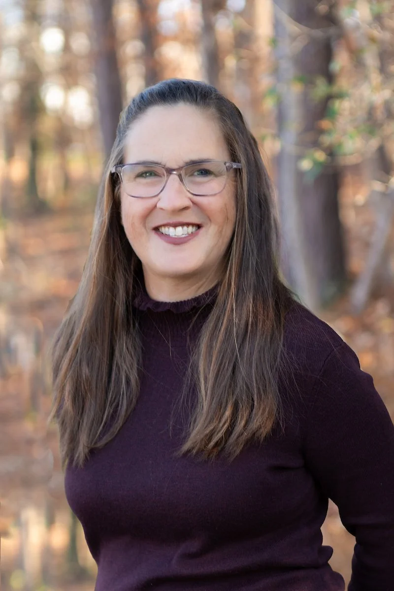 Woman with long brown hair wearing glasses and a burgundy sweater standing in front of an autumn forest. She has a friendly smile.