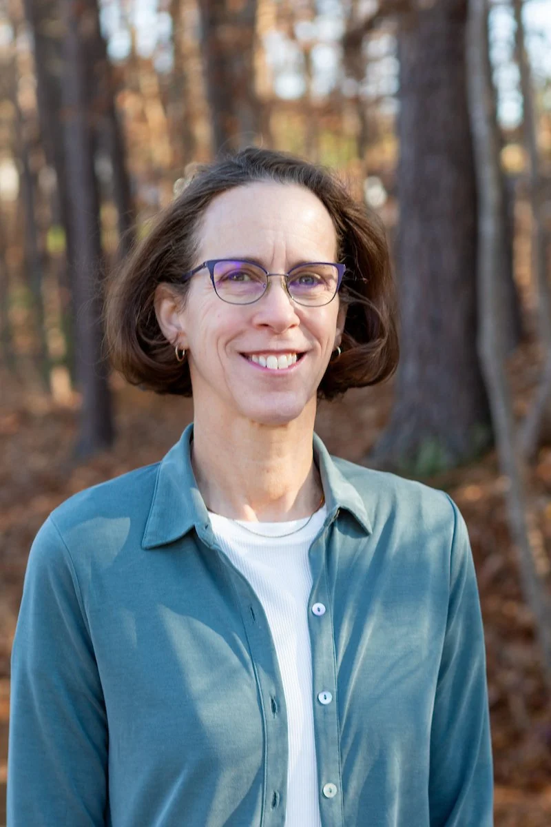 A smiling woman with short brown hair and glasses standing outdoors in front of an autumn forest.