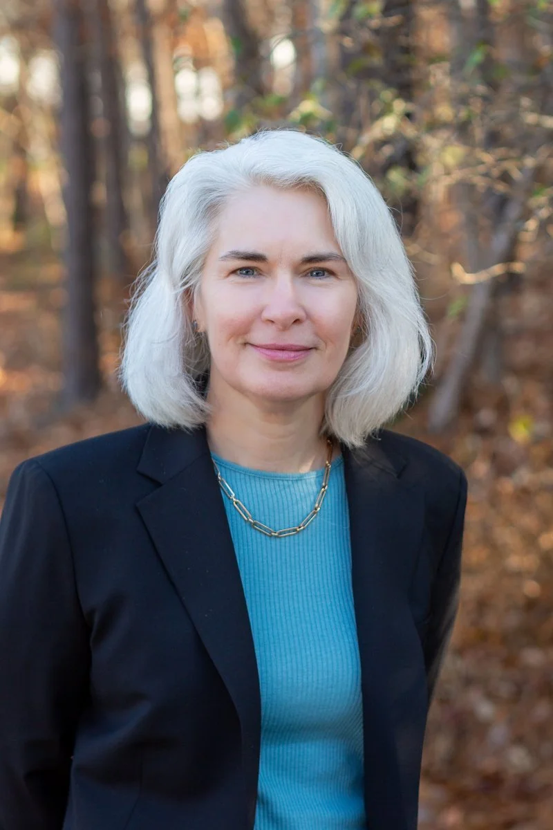 A woman with shoulder length white gray hair in a blue shirt and black blazer smiling warmly, standing in front of an autumn forest background.