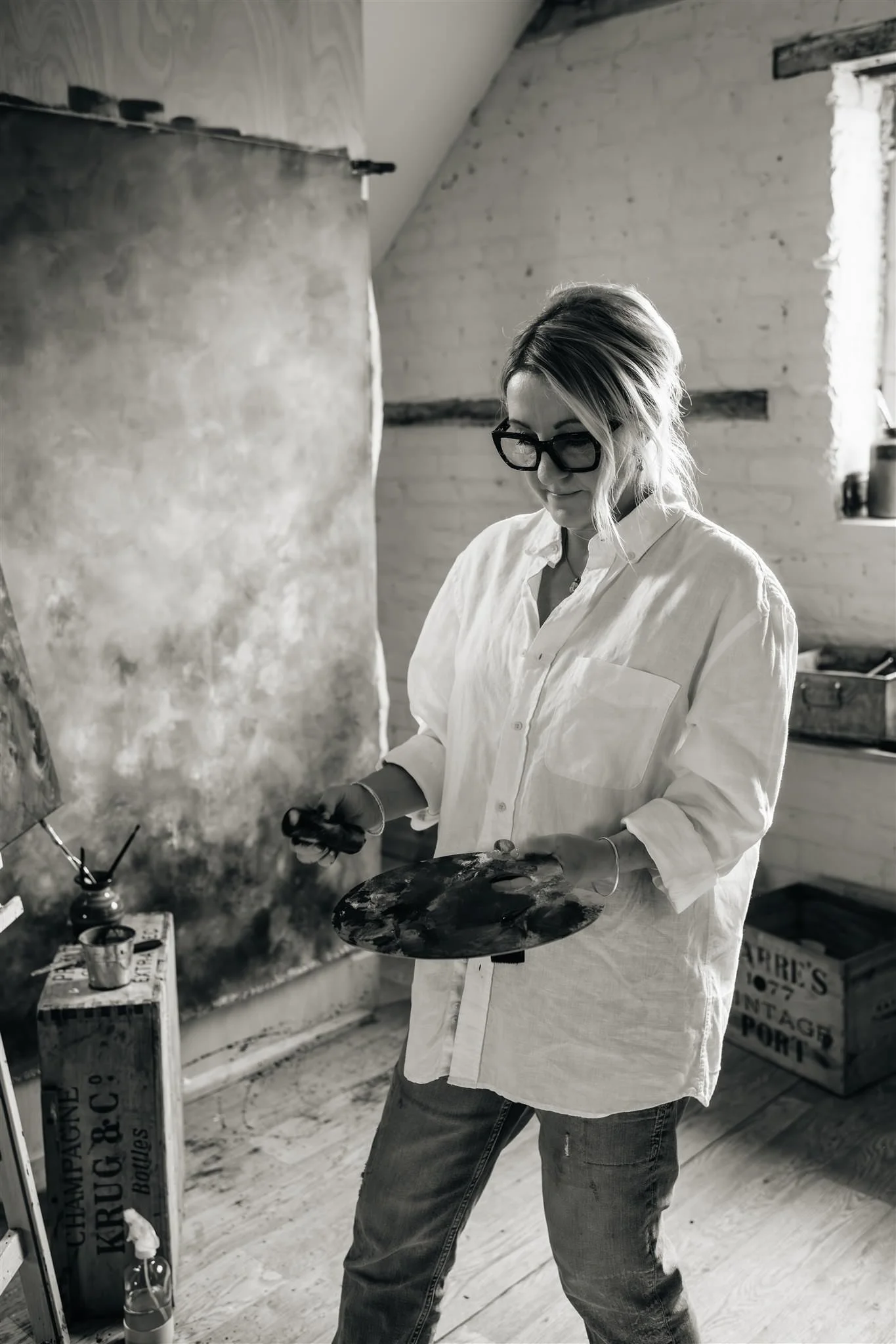 A black and white image of artist Sarah Pitt standing, holding a paint palette, in her studio painting an abstract landscape wearing a pale short and jeans, in her studio with skylights, wooden floor and white walls