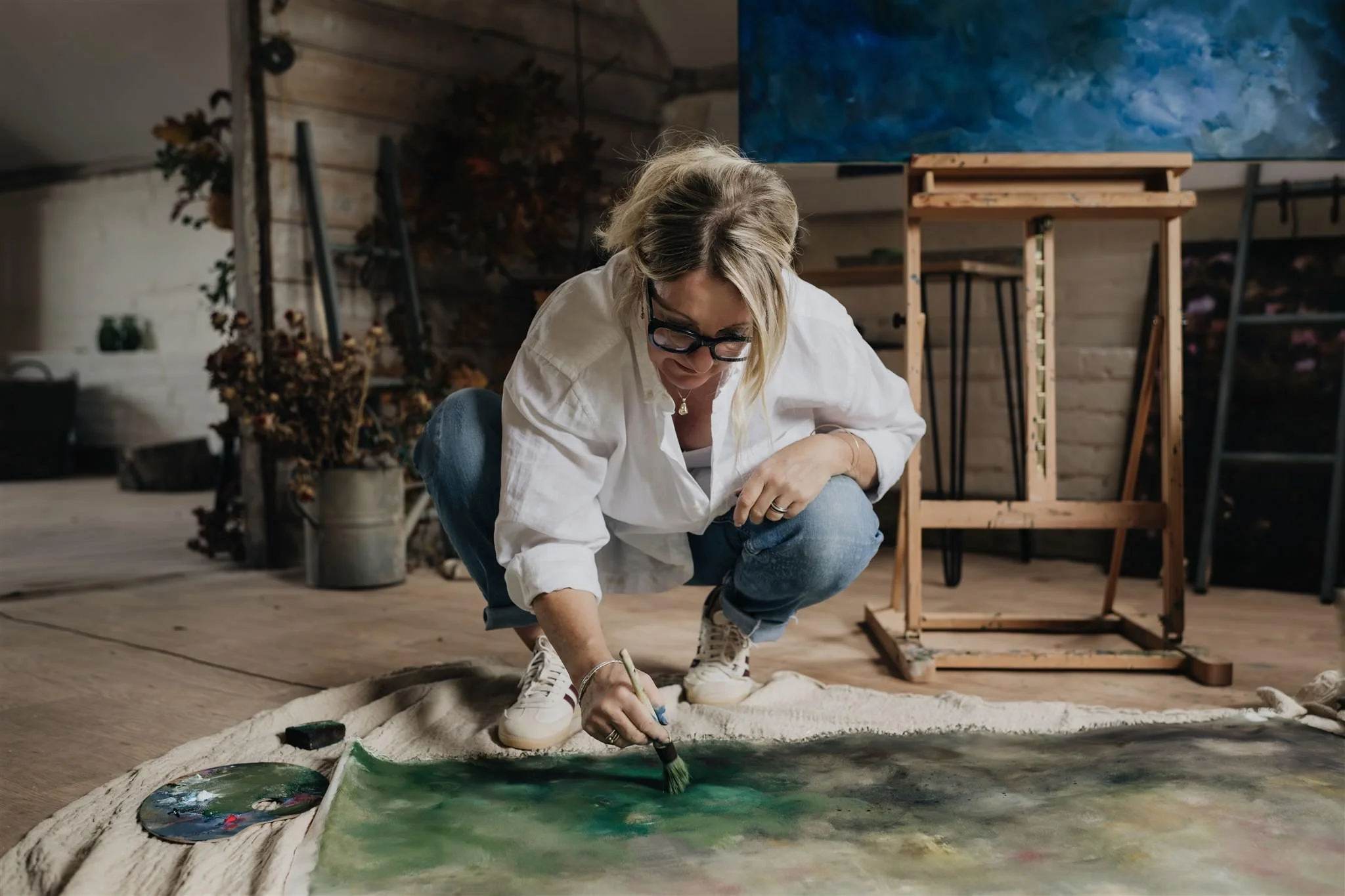 Artist Sarah Pitt crouching on the floor, painting on a large canvas laid out on a cloth. She is using a brush with green paint, wearing glasses and casual clothes in her artist's studio with wooden walls and art supplies around.