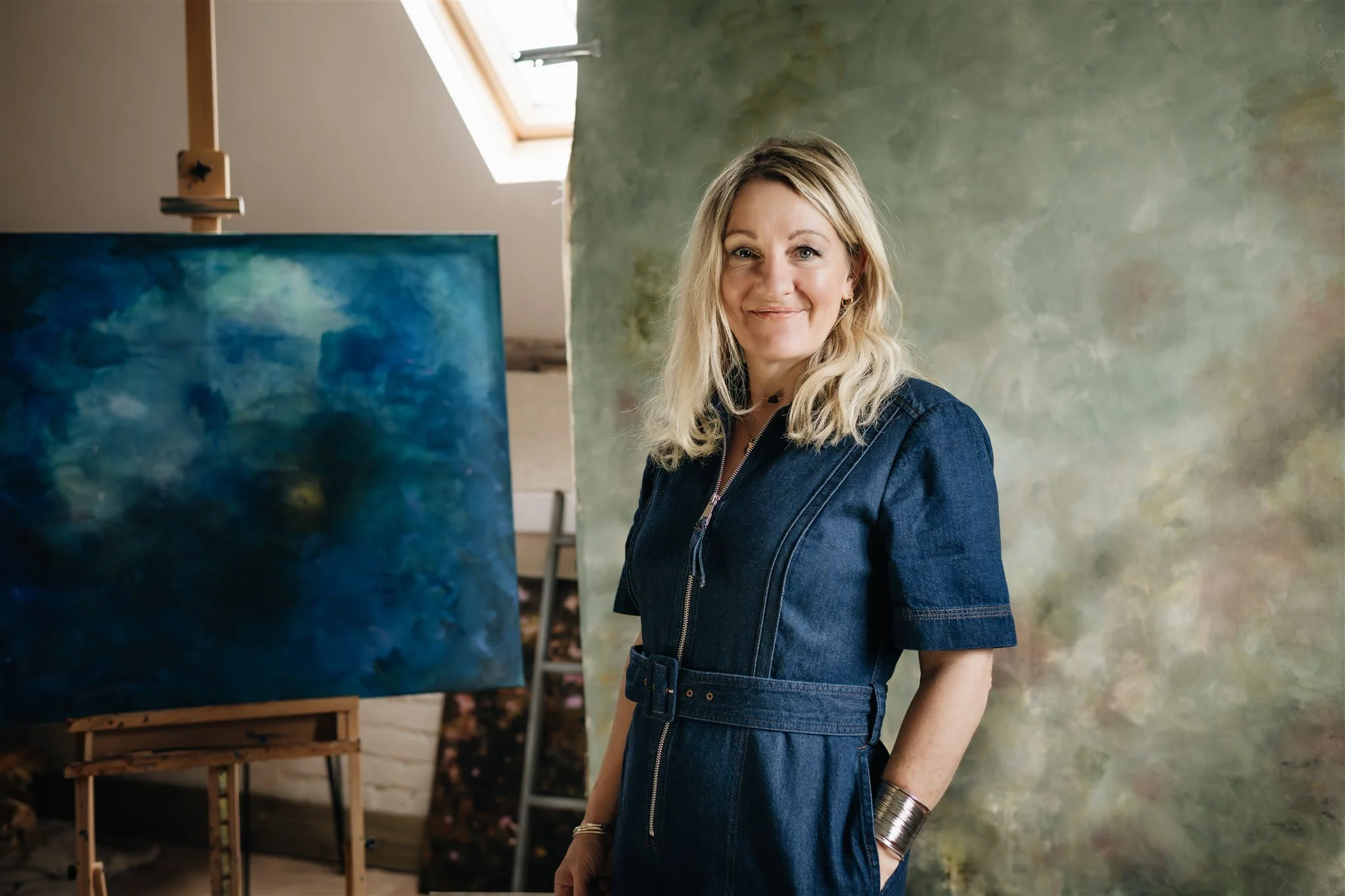 Sarah Pitt an artist with long blonde hair wearing a blue denim jumpsuit stood smiling in her studio in front of her painting