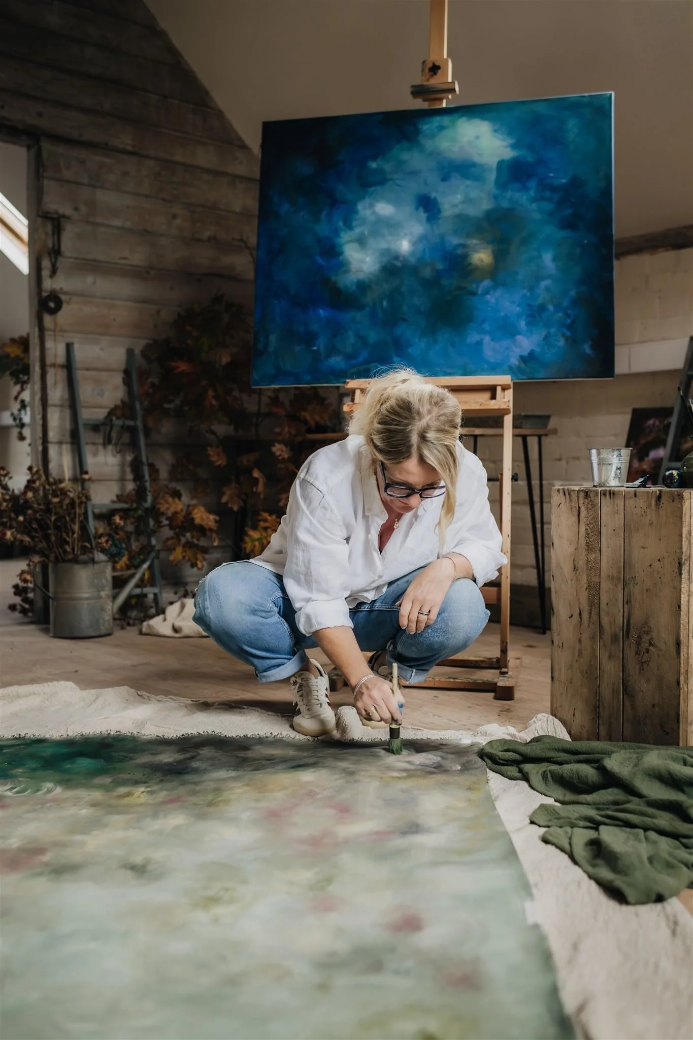 artist wearing awhite shirt and blue jeans crouching over a large hand-painted canvas in her studio.
