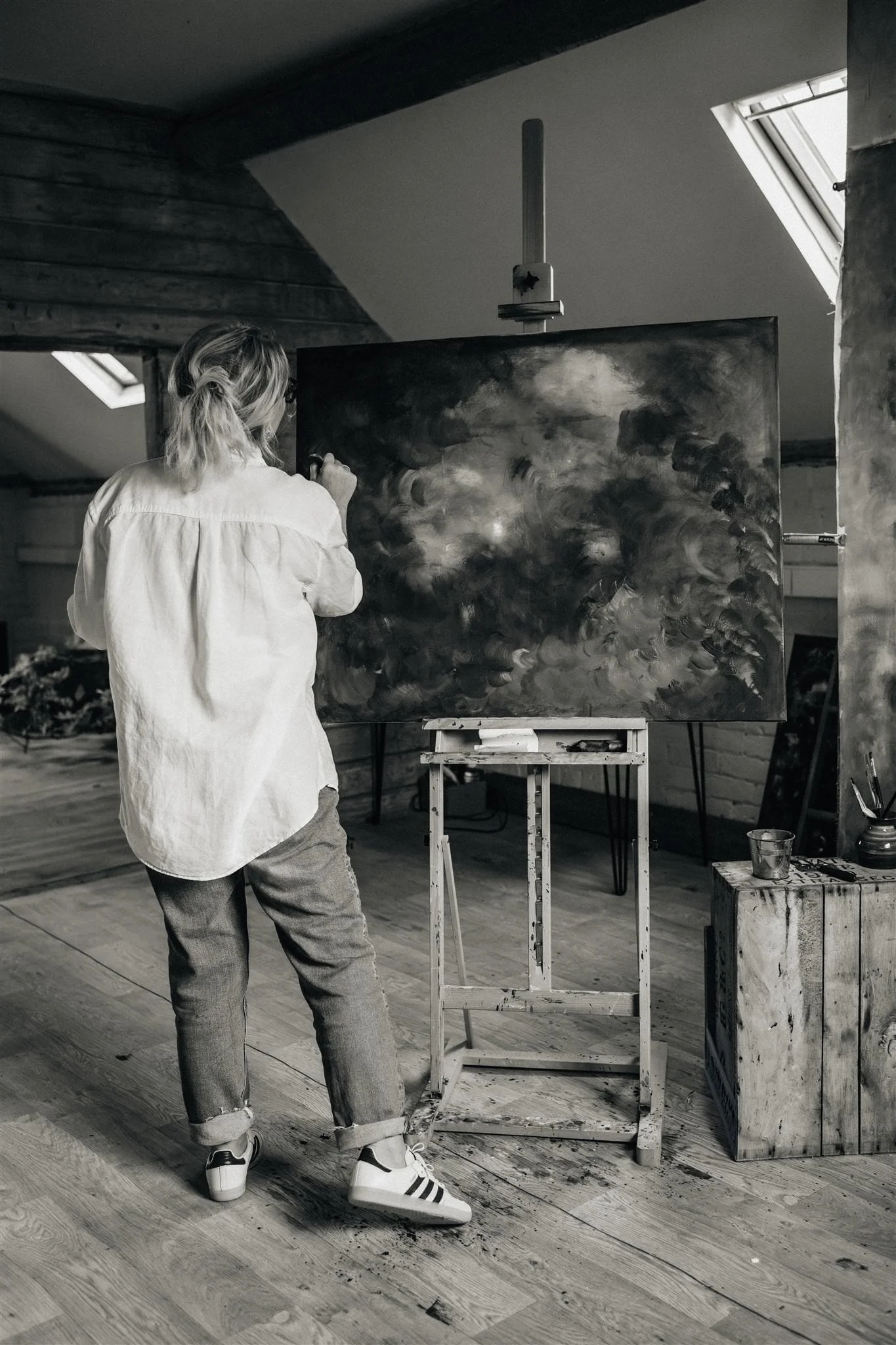A black and white image of artist Sarah Pitt standing, with her back to the viewer, in her studio painting an abstract landscape wearing a pale short and jeans, in her studio with skylights, wooden floor and white walls