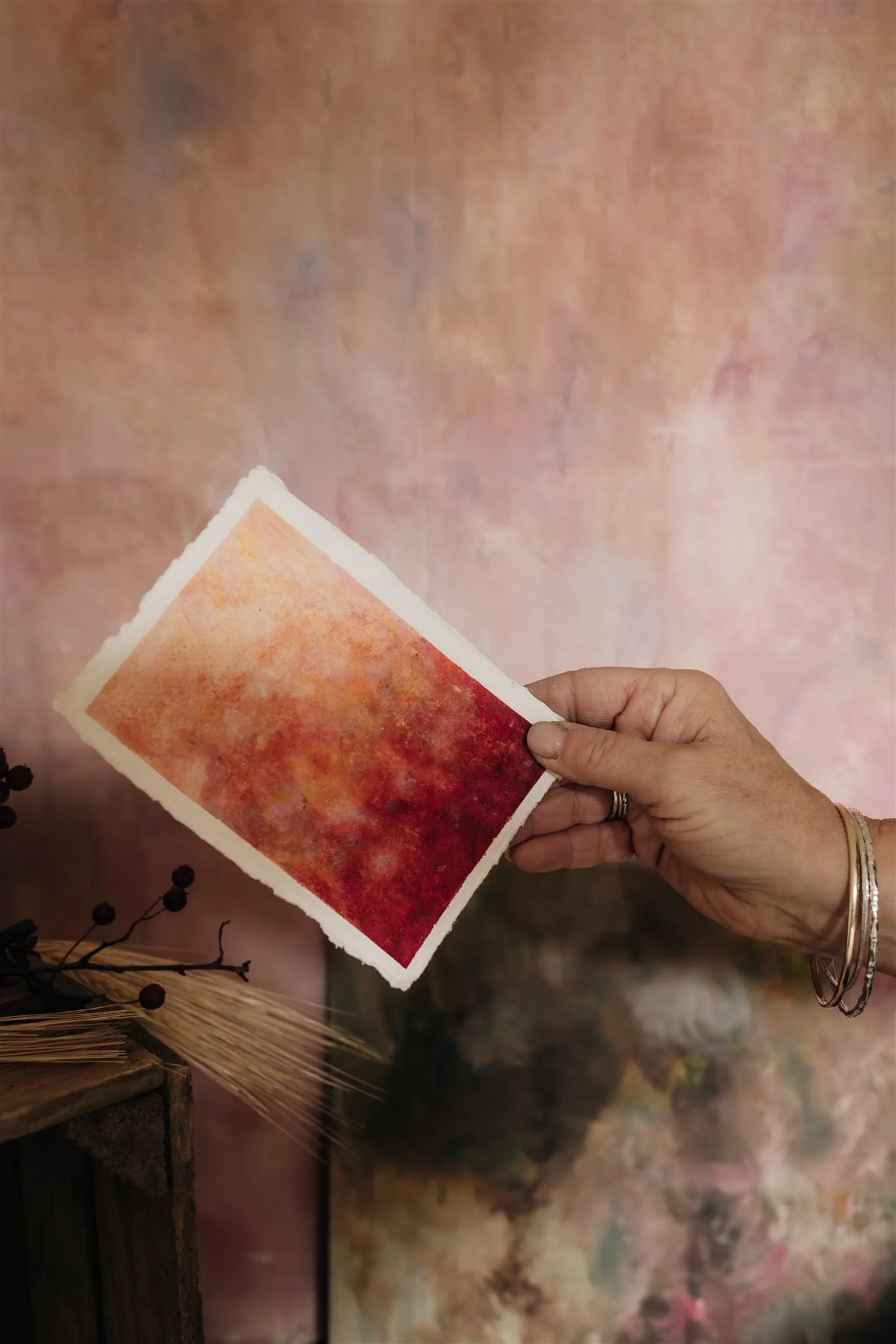 A person's hand holding a small watercolor painting with warm shades of red, orange, and yellow, against a soft, abstract pink and peach background.