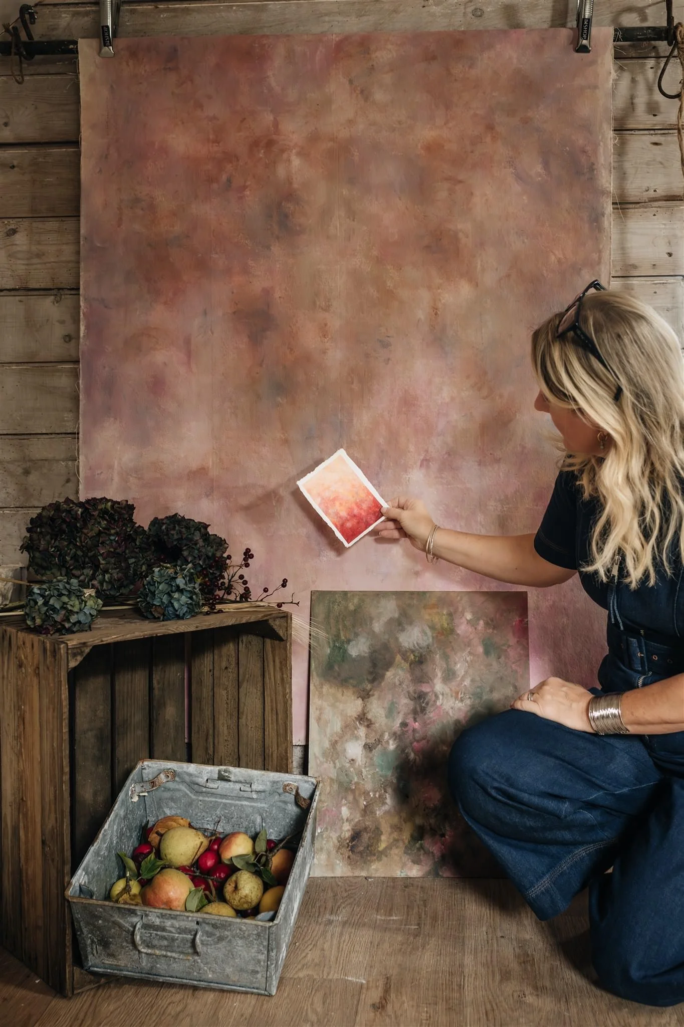 Sarah Pitt of Sarah Pitt Studio sitting on the floor of her artist studio with art supplies, holding a color sample, surrounded by hand-painted backdrop, a wooden crate with apples and pears, and dried flowers, in a rustic art studio.