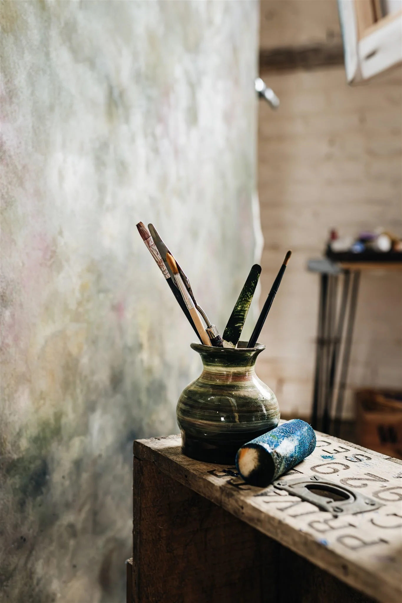 A ceramic vase with paintbrushes and a paint roller placed on a wooden table in art studio of Sarah Pitt.