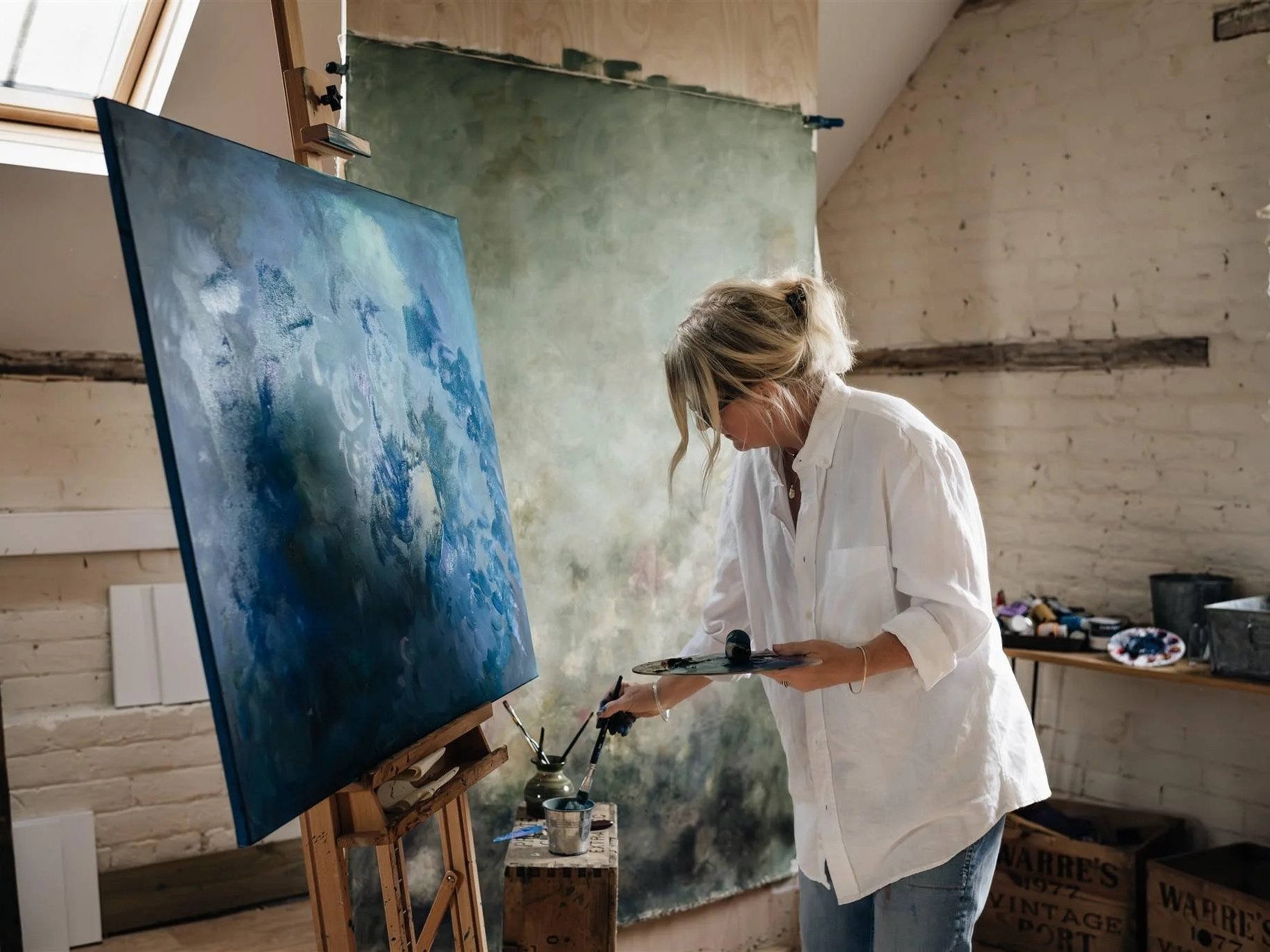 Artist Sarah Pitt in her studio holding a paint palette and paintbrush painting an abstract piece of artwork in blue tones