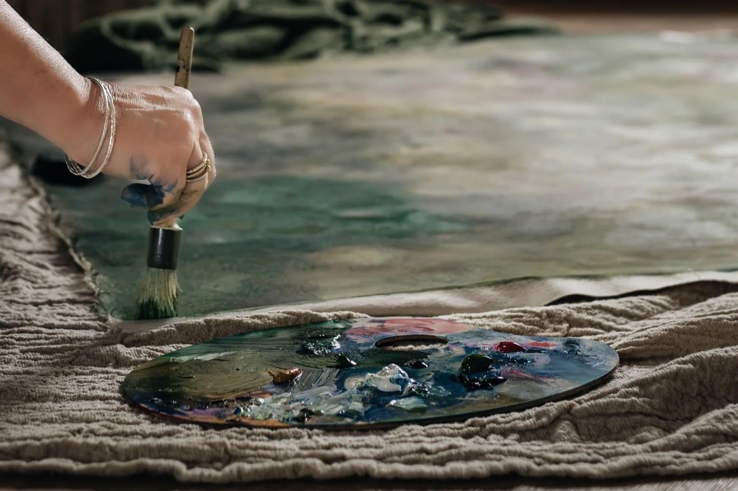 A hand holding a paintbrush is painting on a palette that is resting on a cloth, the artist is painting the canvas o the floor.