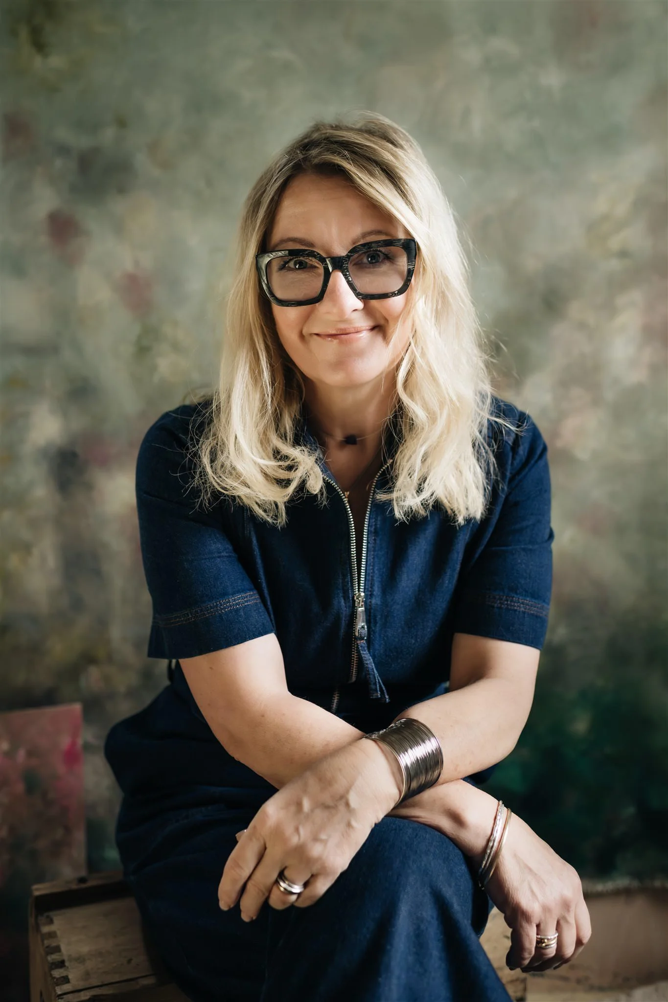 Artist Sarah Pitt with blonde hair and black-rimmed glasses smiling, sitting on a wooden box in front of a painted canvas backdrop.