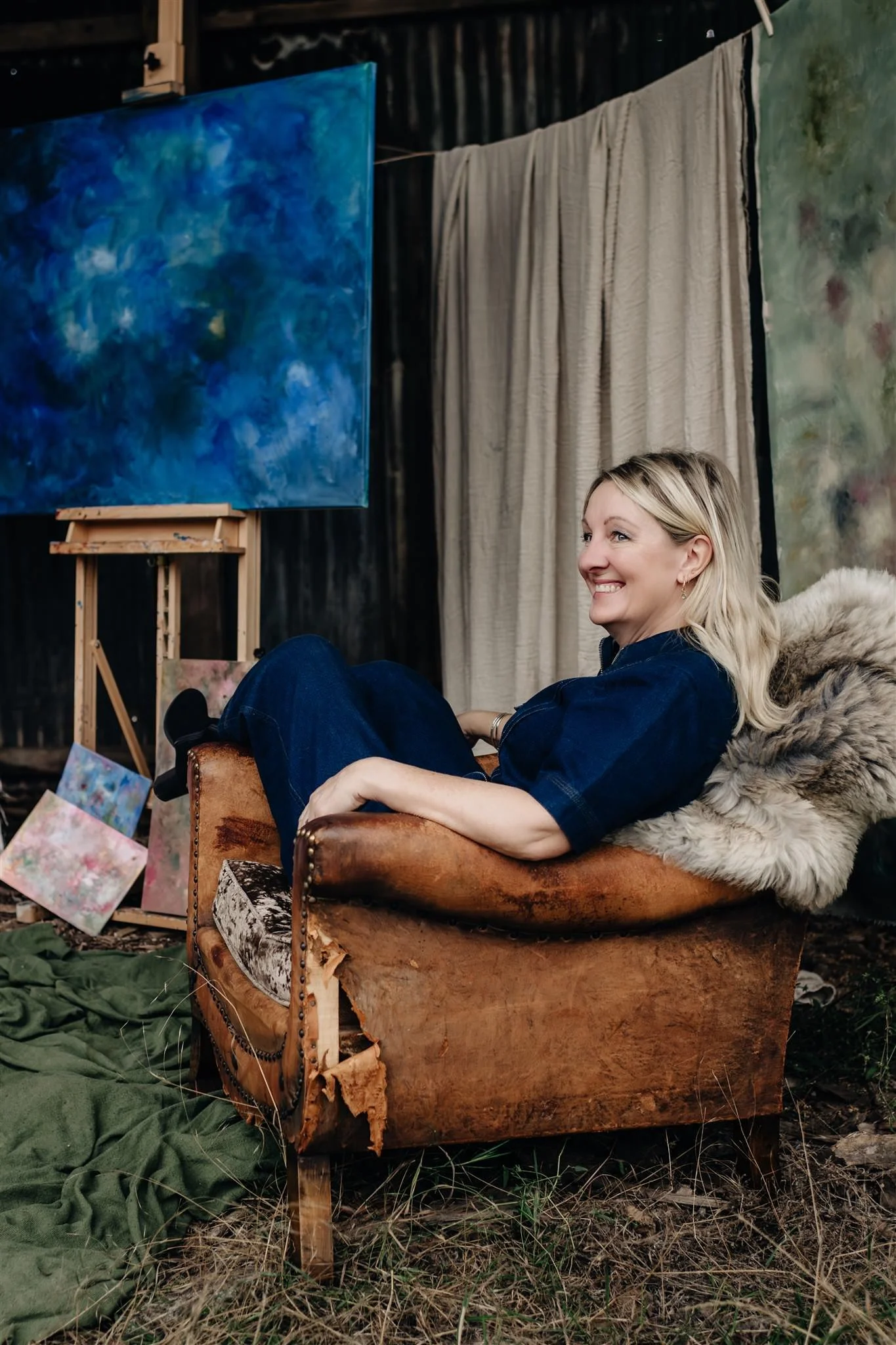 Sarah Pitt, abstract artist, sitting on a worn, tan coloured vintage armchair with a brown sheepskin draped over it, in a derelict barn, smiling and looking to her right, with abstract paintings and artwork and draped fabric in the background.