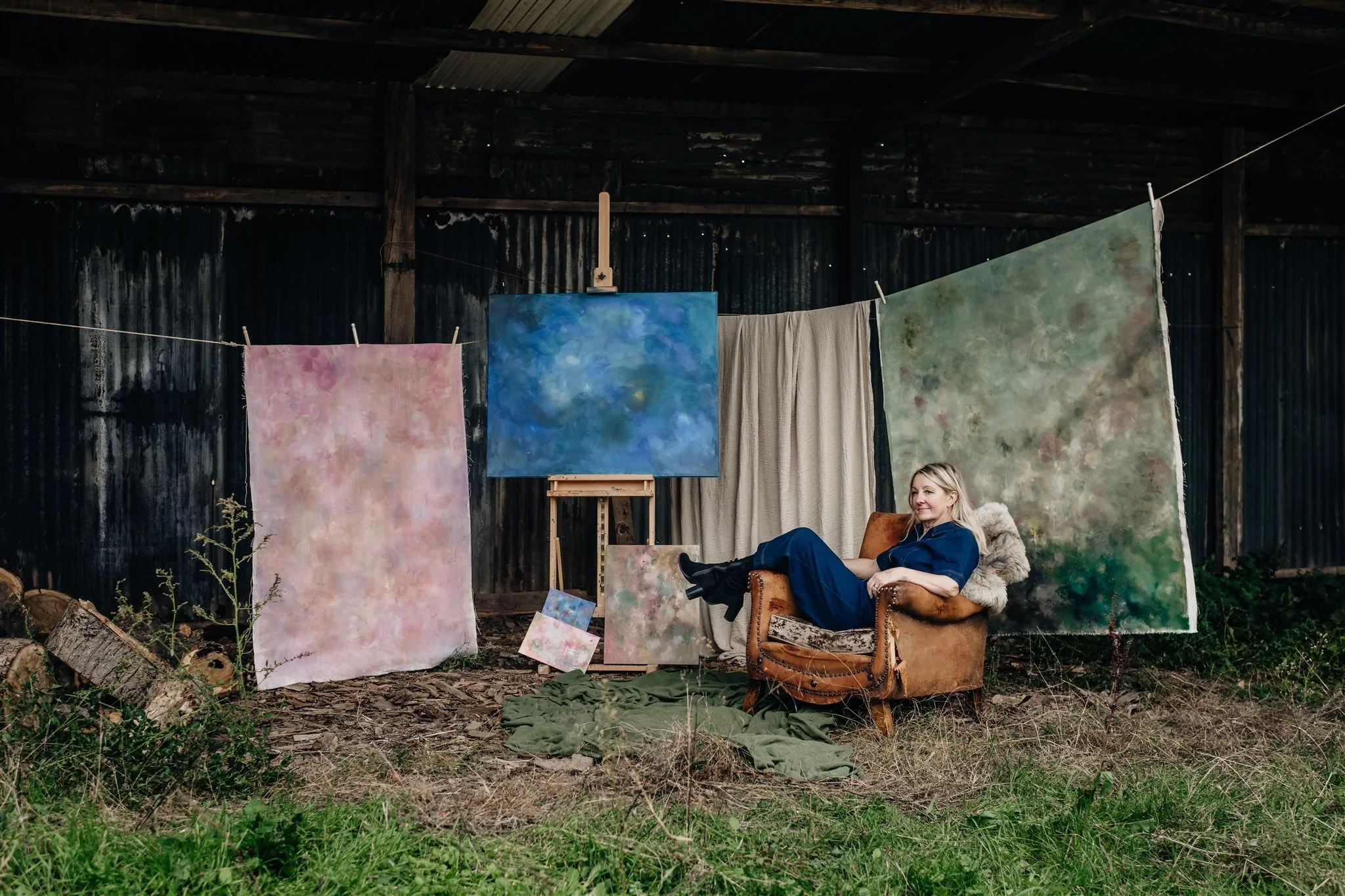 Sarah Pitt sat in an old brown leather chair in a derelict barn next to abstract artwork on a wooden easle and painted canvasses pegged up on a rope line in tones of blue, green and pink