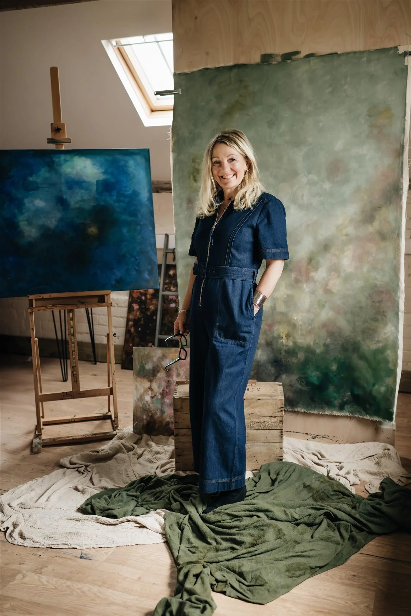 Artist Sarah Pitt standing in her art studio, surrounded by abstract paintings, a green toned hand-painted canvas backdrop, and draped cloths on the floor.