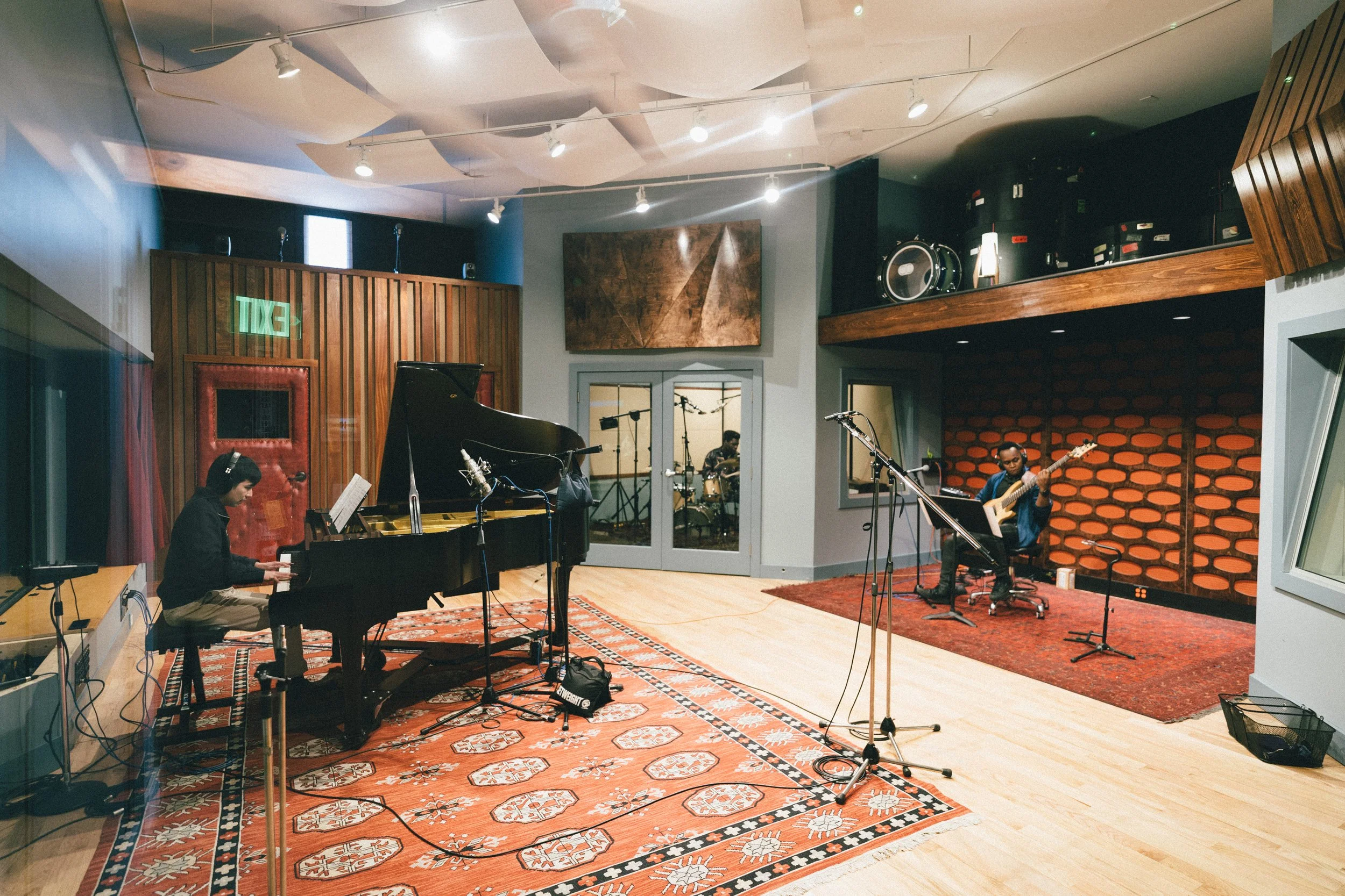 A music recording studio with musicians in a glass-enclosed recording booth. There is a piano on the left side with a musician playing, a drummer in the background, and a guitarist on the right side.