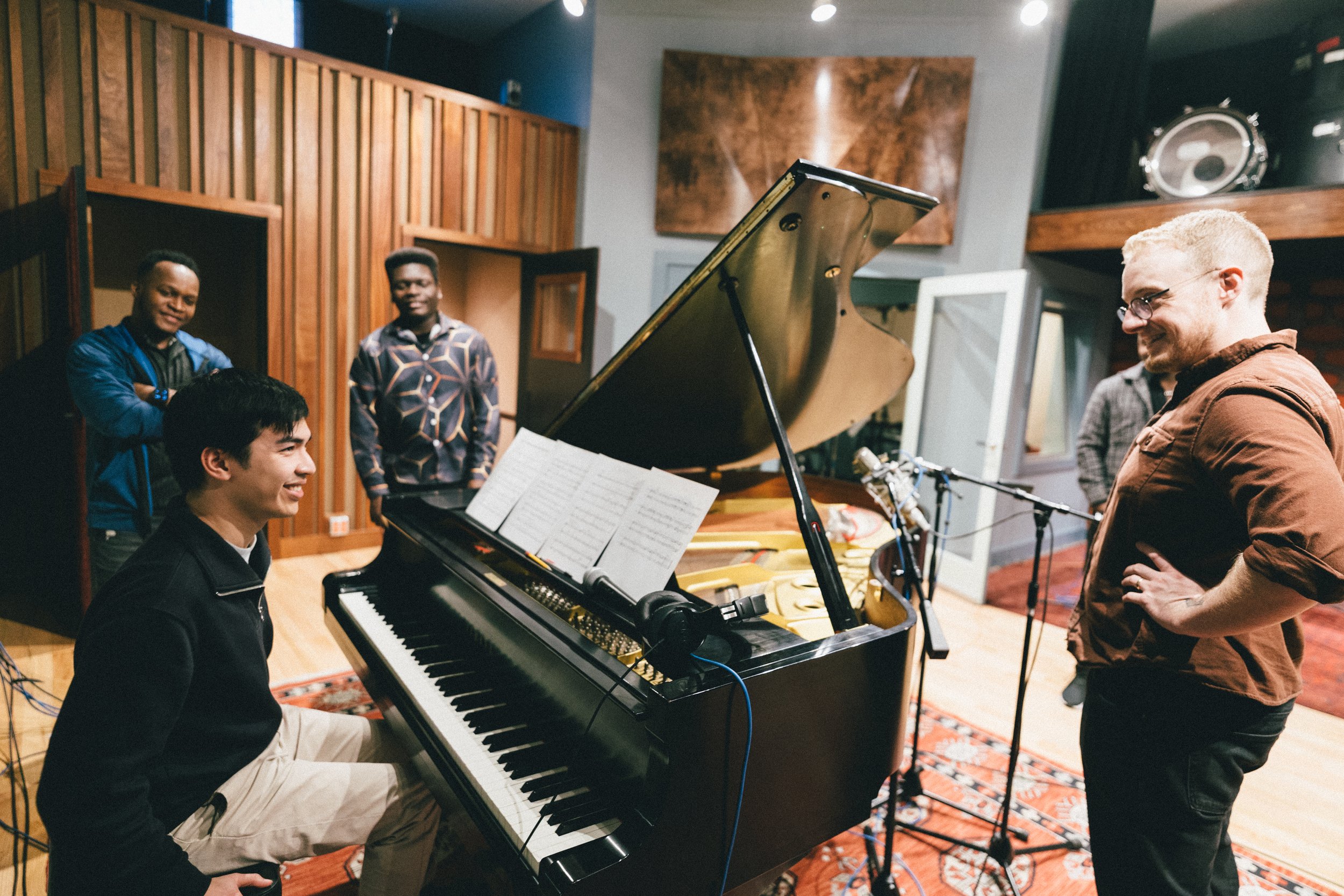 People in a music recording studio. A man playing the piano, a man smiling on the right side, and three men observing and smiling in the background.