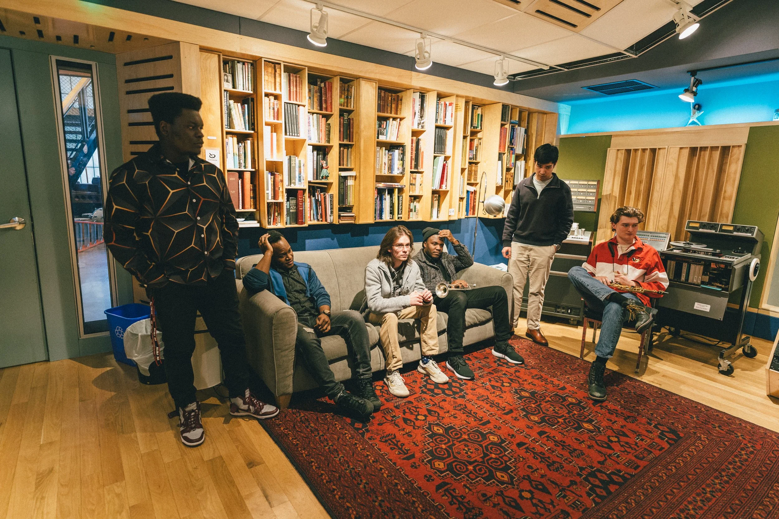 Group of six young people in a music studio with bookshelves, a vintage carpet, and music equipment, some sitting on a sofa and others standing or sitting nearby.