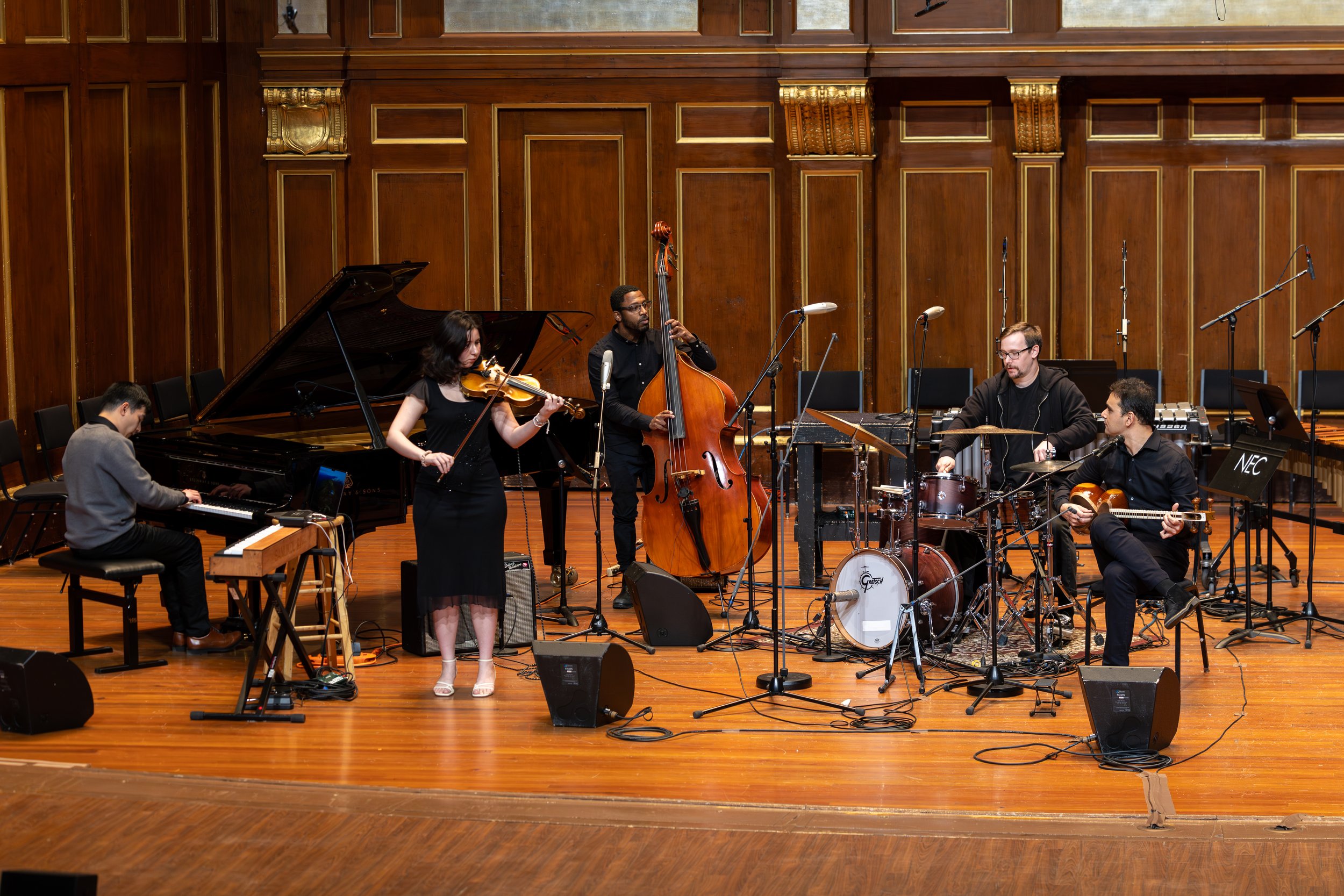 A jazz band performing on stage, featuring a pianist, a violinist, a double bass player, a drummer, and a guitarist in a concert hall with wooden walls.