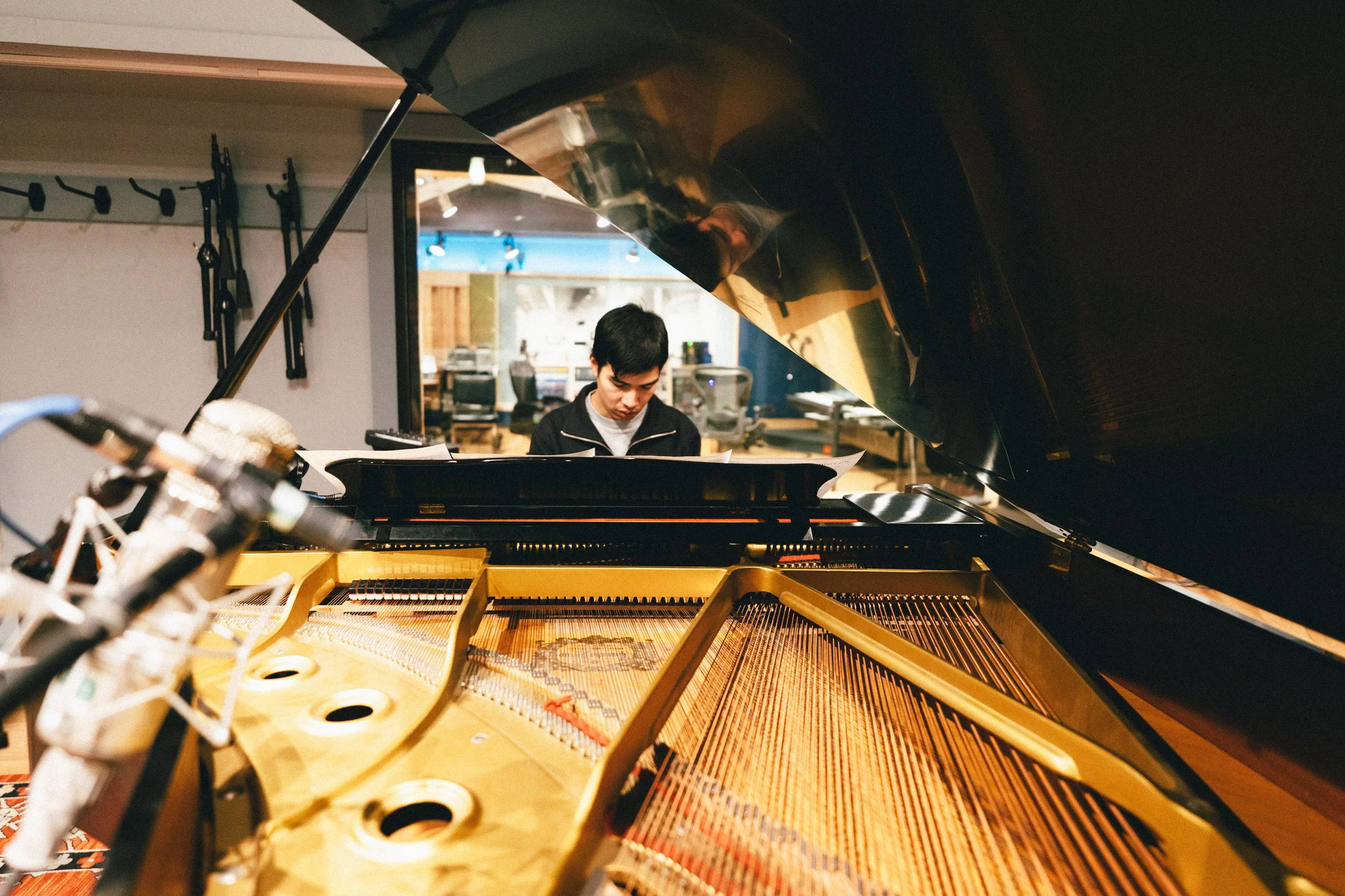 A person sitting at a grand piano, reading sheet music in a recording studio with music equipment and monitors in the background.