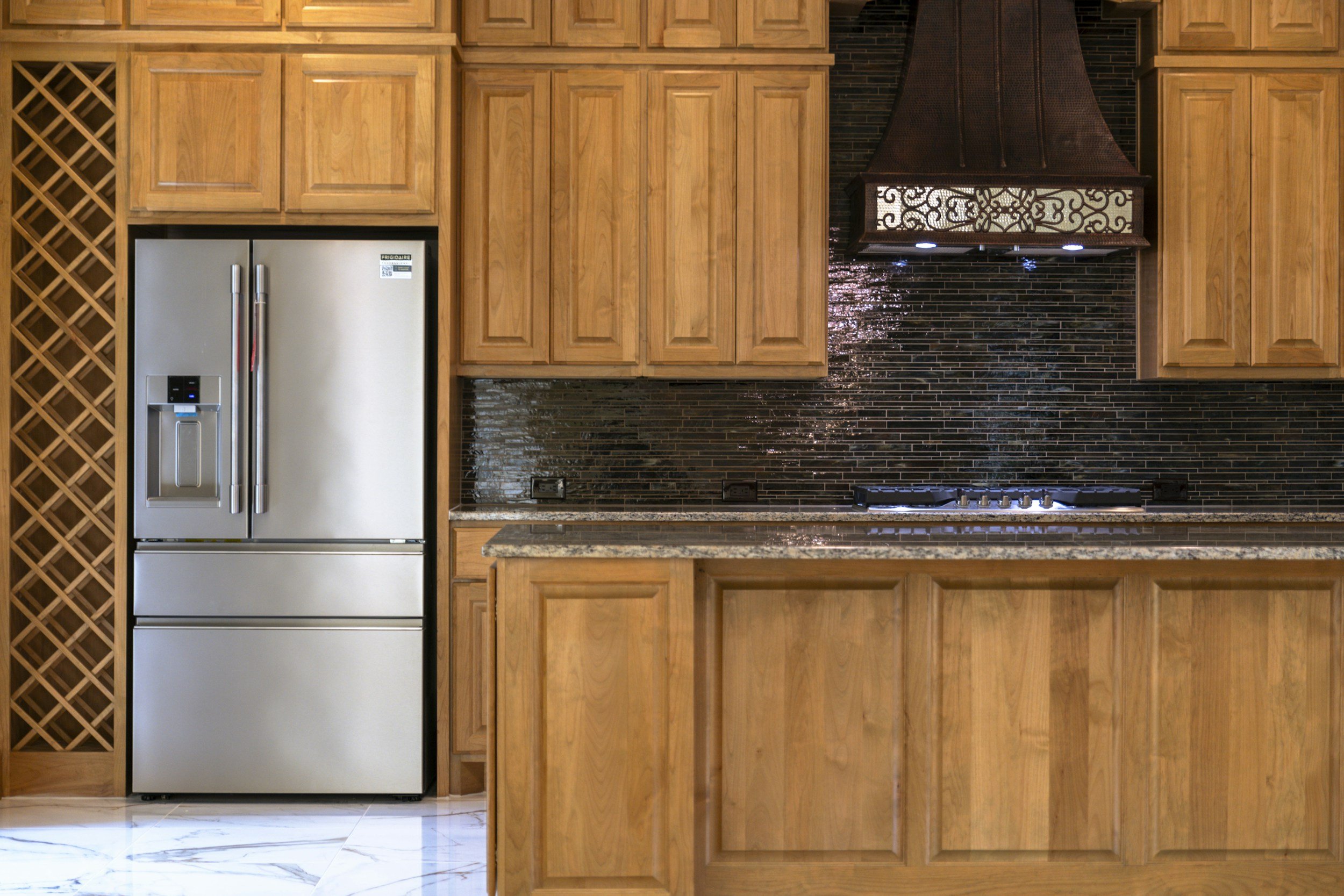 Kitchen with wooden cabinets, black tile backsplash, stainless steel refrigerator, and a stove with a decorative range hood.