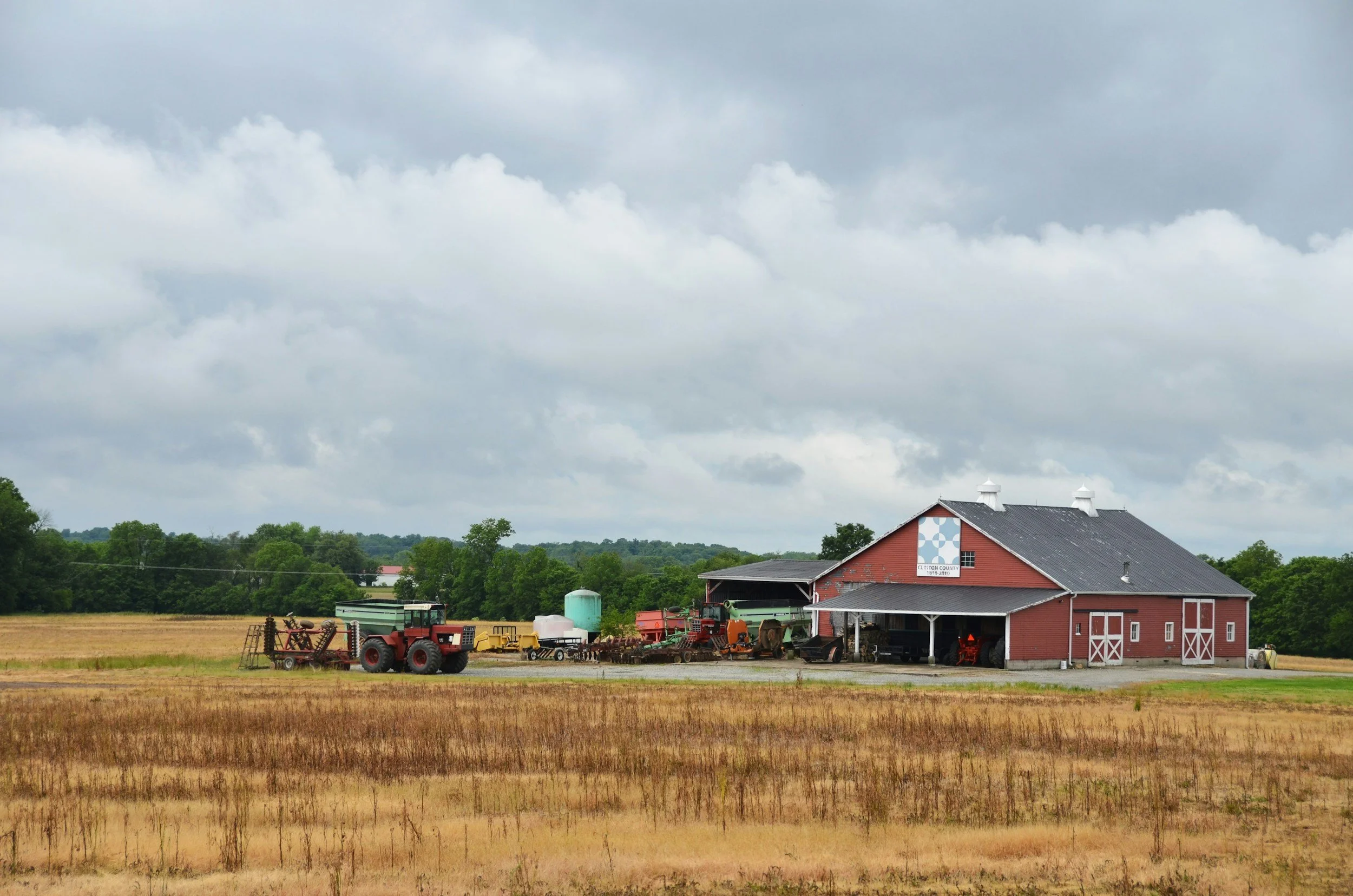 A red barn on a farm surrounded by farmland, with farm equipment and machinery, under an overcast sky.