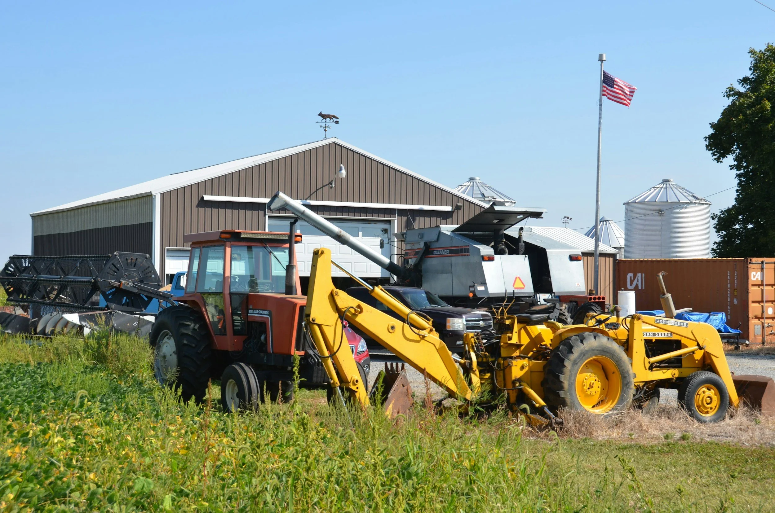 Farmyard scene with tractors, a barn, grain silos, an American flag, and farming equipment.