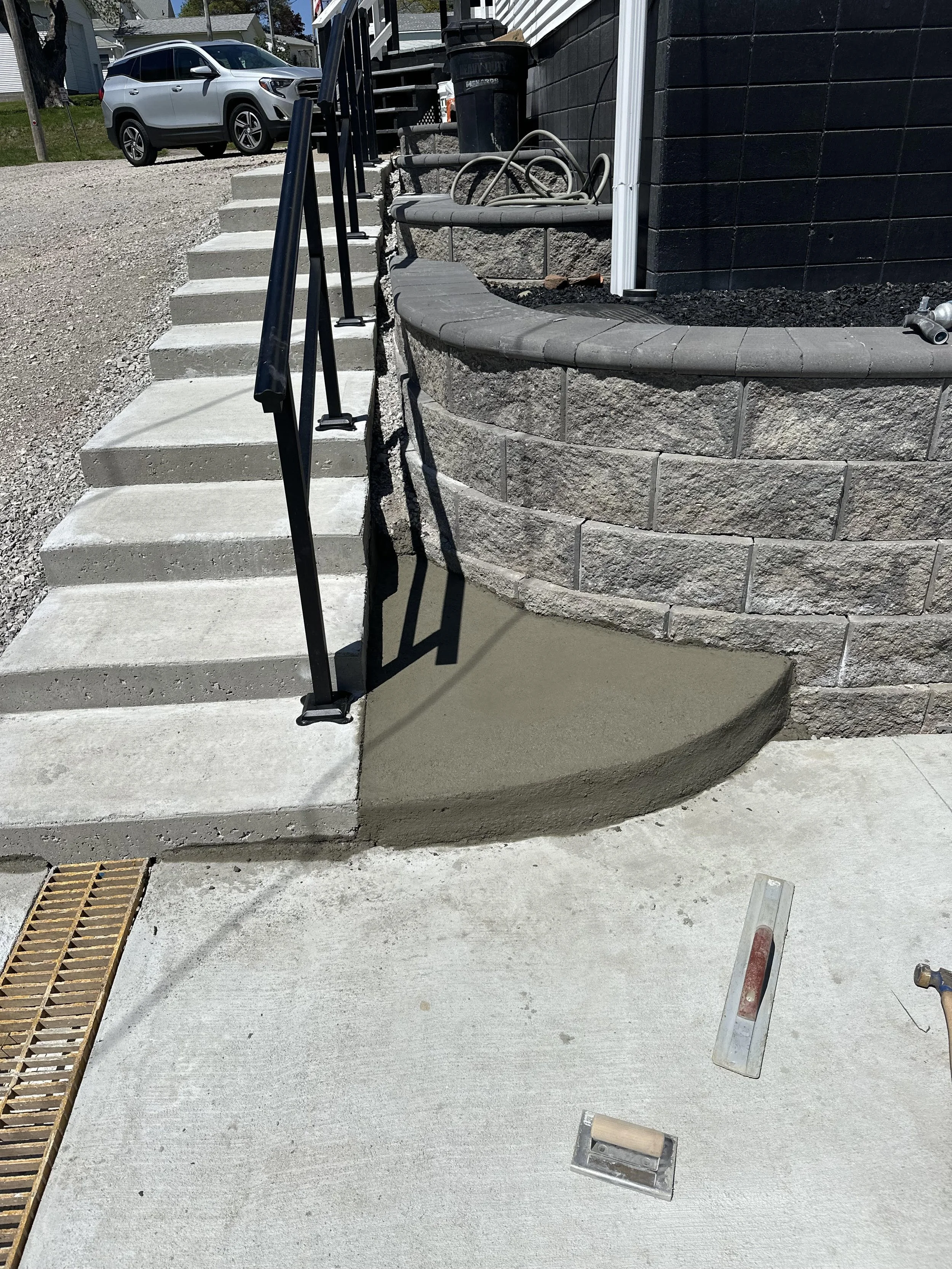Construction site showing new concrete steps, metal handrails, and a curved concrete pad in front of a stone retaining wall next to a black house siding.